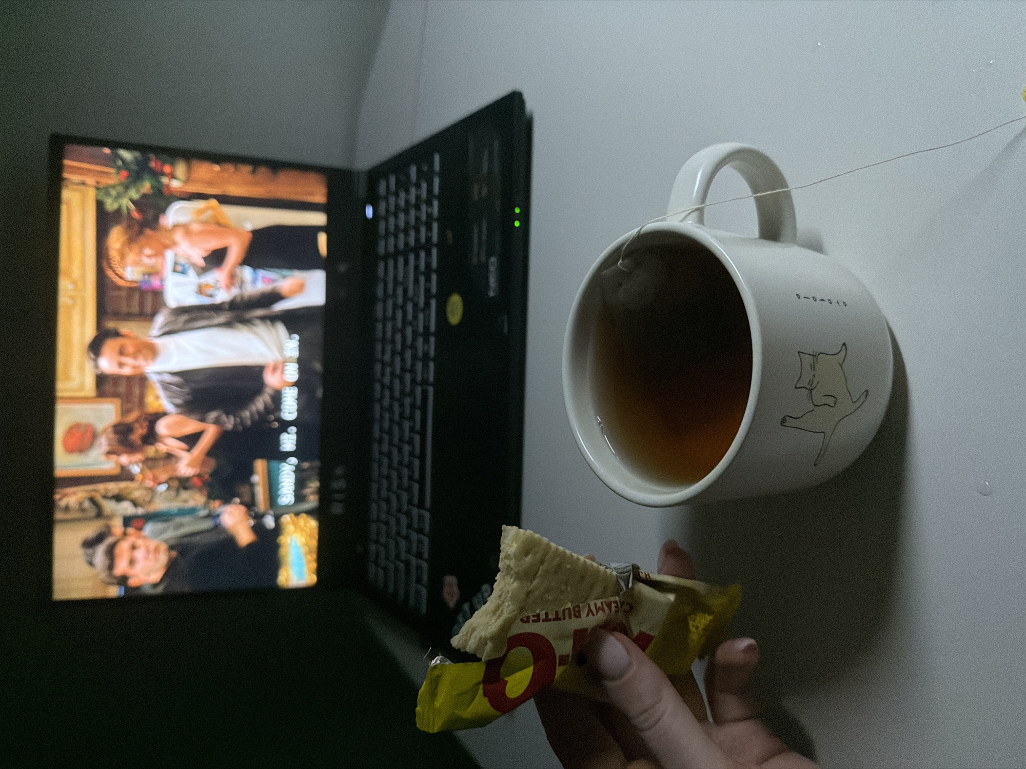 A person's hand, with lightly manicured nails, holds a partially eaten snack bar with yellow packaging and red lettering that reads "CREAMY BUTTER". In front of the hand is a white mug with a drawing of a cat on it, filled with amber liquid and a tea bag string hanging out. Behind the mug and snack bar, a laptop is open, displaying a scene from a television show, likely "Friends", with actors Chandler Bing, Monica Geller, and Rachel Green visible. The laptop is on a white surface, with a wall in the background that appears to be a muted gray. The scene is dimly lit, suggesting an indoor setting, possibly a living room or bedroom in Dongducheon-si, South Korea.