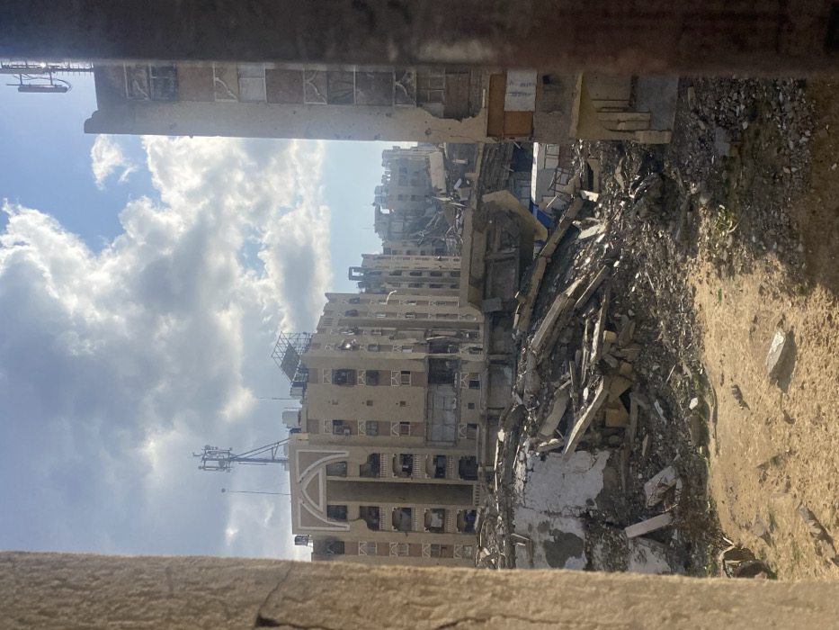The image displays an urban landscape in Khan Yunis, Palestinian Territory, viewed through an opening framed by two rough vertical structures, likely walls or pillars. The foreground consists of dry, light brown earth mixed with significant amounts of concrete rubble and debris. In the midground, an extensive pile of collapsed building materials, including concrete slabs, twisted rebar, and fractured elements, dominates the scene, indicative of widespread destruction. Further back, a multi-story, beige-colored apartment building stands prominently, exhibiting considerable damage such as broken windows, missing facade sections, and scorch marks. A communications tower with multiple antennae is situated on its roof. To the right, another multi-story building, partially obscured, also shows severe damage, including broken windows and sections of its exterior walls appearing heavily impacted. In the background, additional structures appear heavily damaged or partially collapsed, contributing to the scene of devastation. The sky is partly cloudy with patches of blue, under bright daylight conditions. No individuals are visible in the scene.