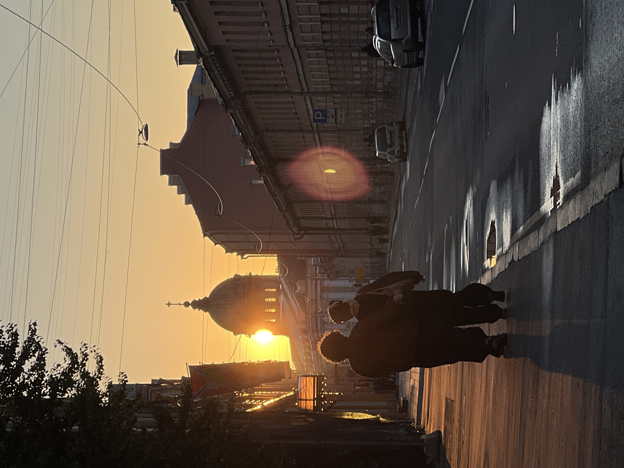 The image captures a sunset in Saint Petersburg, Russia, casting a warm, golden glow over the scene. The sun is low on the horizon, peeking through the dome of a grand building, possibly a church or government structure. The warm light reflects off the wet pavement, creating elongated, shimmering streaks.
In the foreground, two figures are walking away from the camera, their silhouettes dark against the bright sunset. They appear to be engaged in conversation or simply enjoying the evening stroll. Behind them, a street runs alongside a row of historic buildings, their facades detailed and ornate. A few cars are visible on the street, and there's a "P" sign, indicating a parking area. The sky is a soft gradient of orange and yellow, indicative of a clear sunset.
The overall mood is serene and contemplative, with the warm light and quiet street scene evoking a sense of peace. The architecture suggests a European city, and the context provided points specifically to Saint Petersburg, a city known for its historical buildings and beautiful sunsets. The wet pavement might indicate recent rain or a morning dew that has not yet evaporated. The image is shot vertically, with the sunset and dome dominating the left side of the frame, and the street and figures on the right.