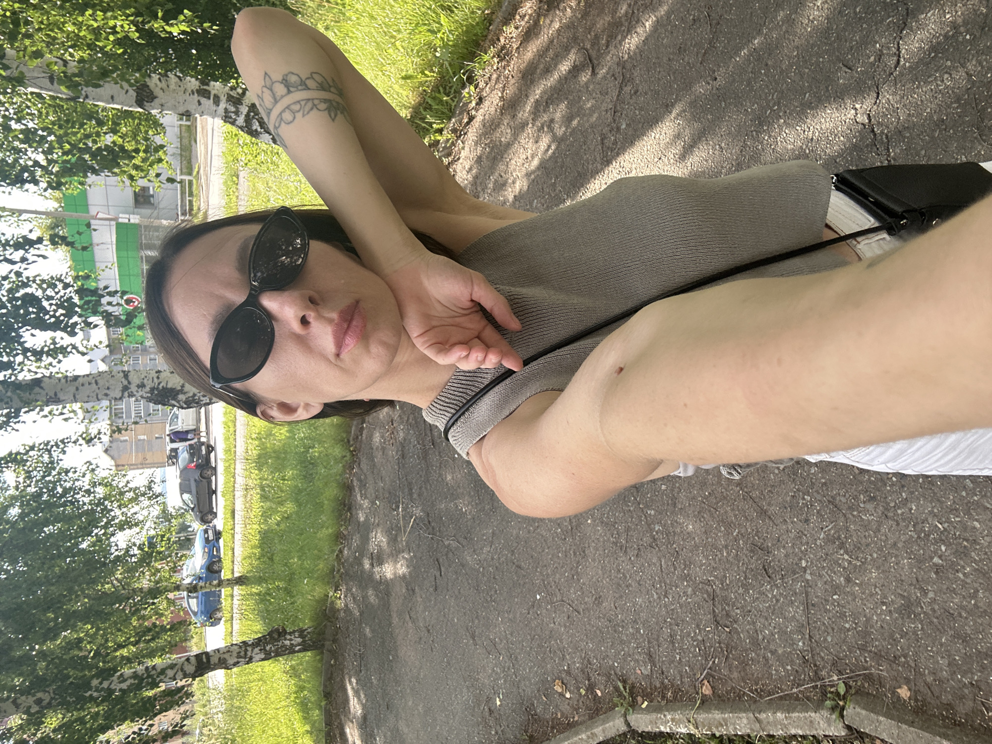A woman with sunglasses and a tattoo on her arm is taking a selfie outdoors. She is wearing a light grey tank top and white pants. A black purse is visible across her body. The background shows birch trees, a street with cars, and buildings. The scene appears to be a residential area in the city of Votkinsk, Russia, likely during daylight hours.