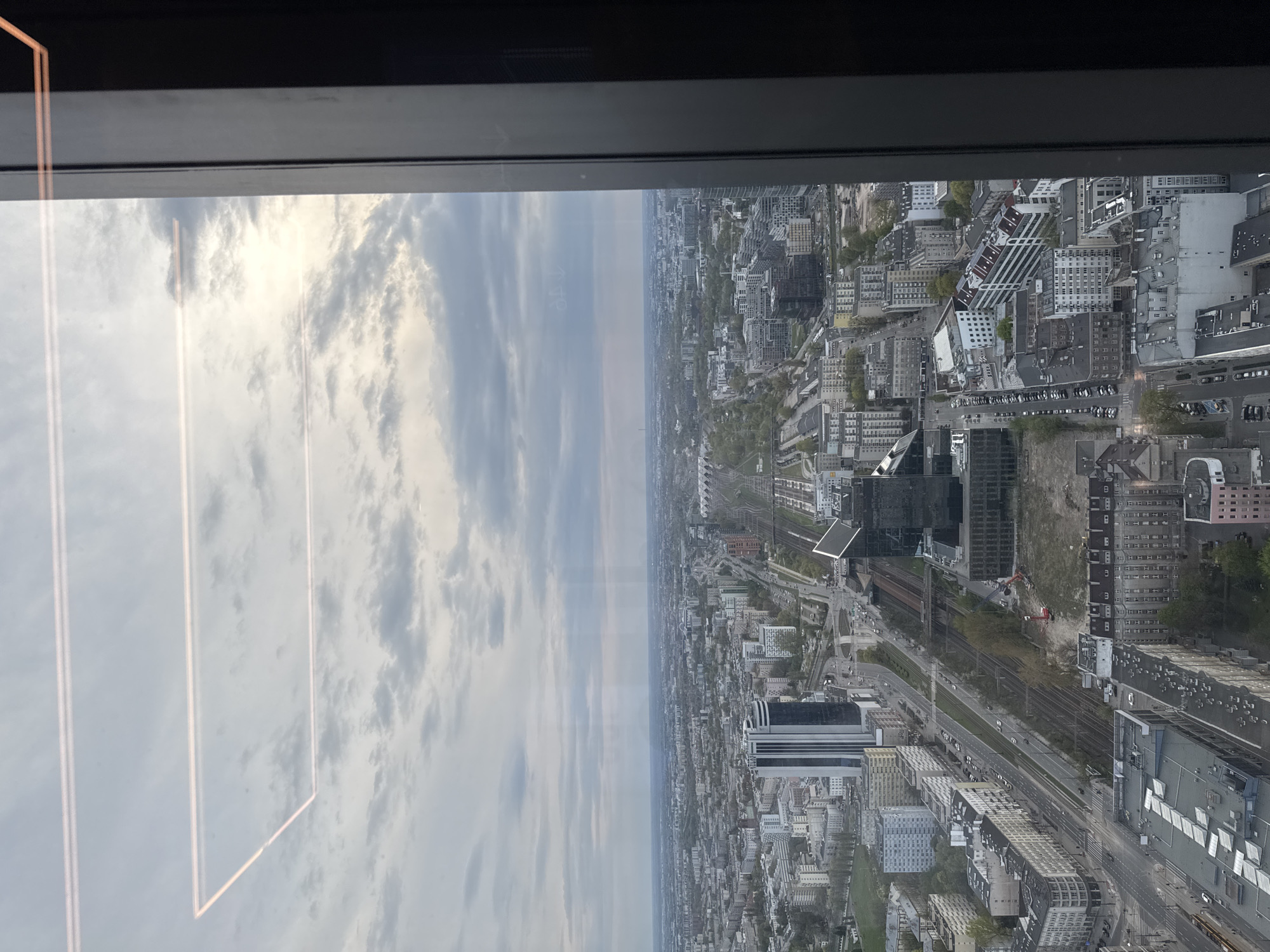 The image shows a high-angle view of Warsaw, Poland, as seen through a window. The sky is cloudy. The cityscape includes numerous buildings of varying heights and architectural styles. A railway line runs through the city. A construction site with a red crane is visible. The window frame is visible at the top of the image and the side of the window is seen on the left side of the image.