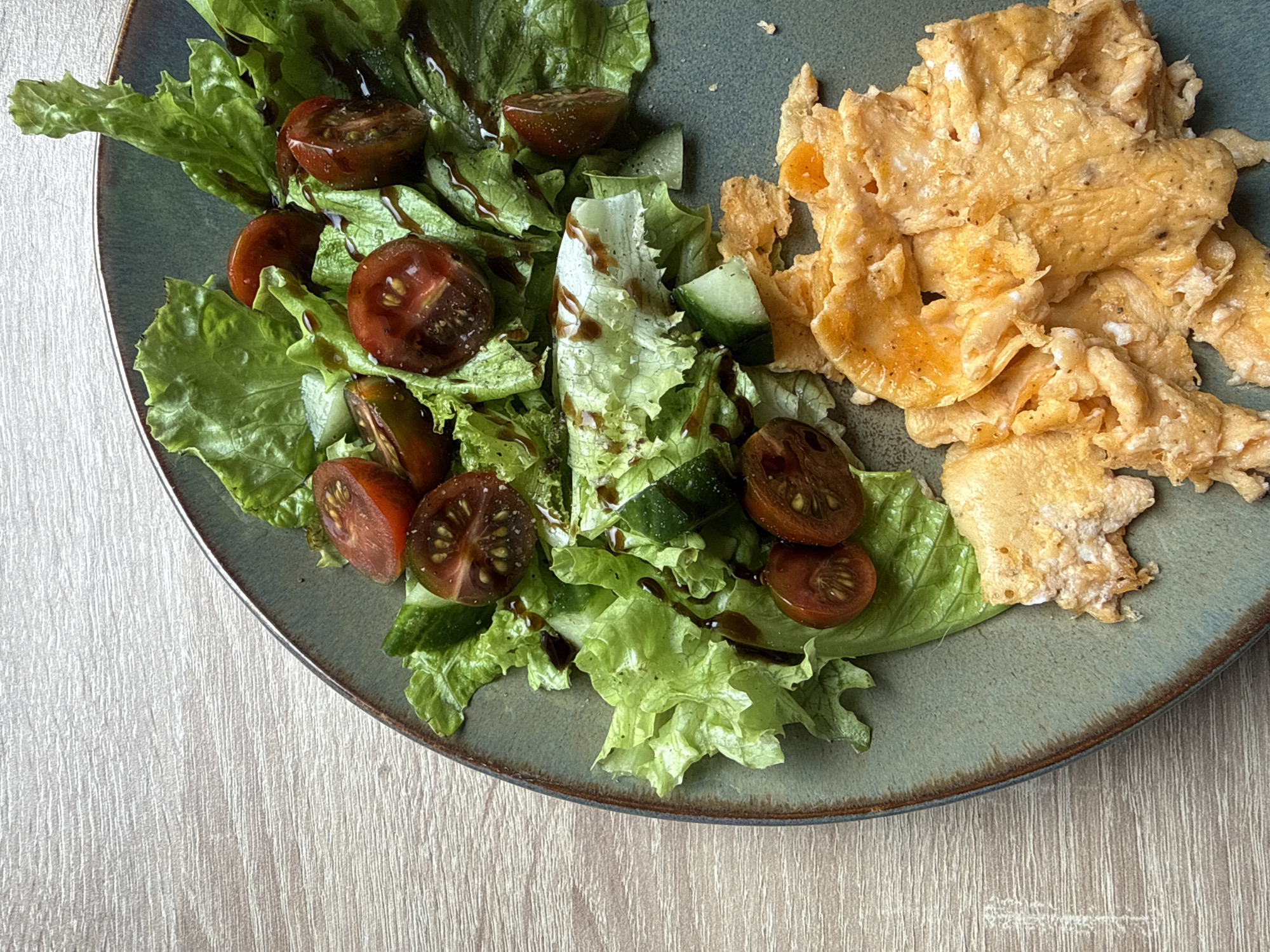 A high-angle shot shows a green plate containing a salad and scrambled eggs. The salad is composed of lettuce, halved dark tomatoes, cucumber, and a dark drizzle (likely balsamic glaze). The scrambled eggs are light yellow and slightly browned. The plate rests on a light-colored, wood-grain surface. There are no people visible. The location is not discernible from the image content.