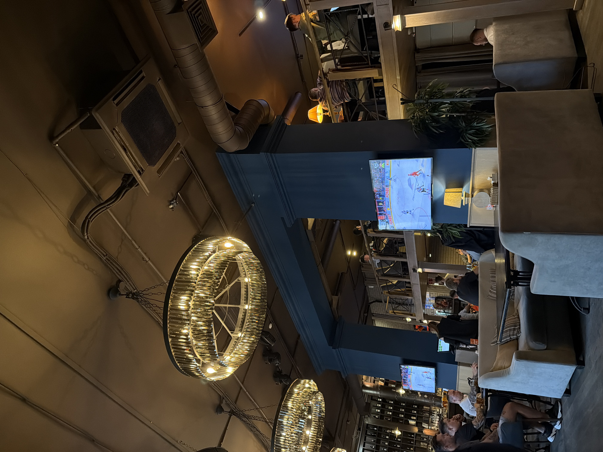 The image shows the interior of a bar or restaurant. There are several people sitting at tables and couches, appearing to be watching a hockey game on television screens mounted on pillars. There are numerous bottles displayed behind the seating area. Above, there are large circular chandeliers and industrial-looking pipes and air conditioning units on the ceiling. The location is Kazan, Russia, as indicated by the FONBET logo on the television screen, a Russian betting company.