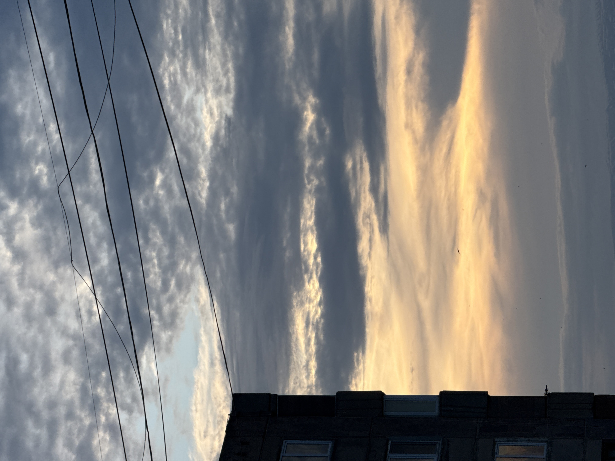 A tall, dark, concrete building stands to the left of the frame, its facade marked by rows of windows and projecting balconies. The sky above is filled with clouds, ranging from light grey and white to darker, stormier hues. A band of warm, golden light illuminates the horizon, suggesting either sunrise or sunset. Several electrical wires are strung across the upper portion of the image, cutting through the cloudy sky. A small bird is visible in flight against the bright horizon. The scene appears to be an urban environment, likely in Abovyan, Armenia, given the architectural style of the building and the characteristic cloud formations often seen in the region.