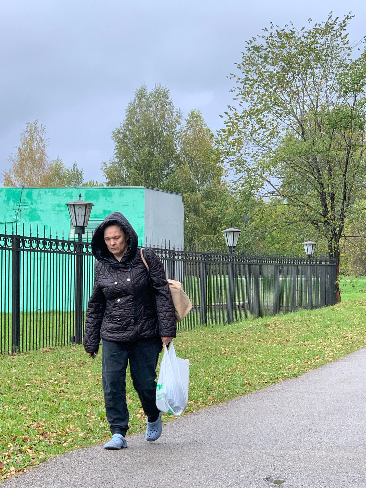A woman walks on a paved path next to a black metal fence. She is wearing a black hooded jacket, dark pants, and blue clogs. She is carrying a beige bag slung over her shoulder and a white plastic bag in her right hand. Behind the fence is a grassy area with fallen leaves and trees. A light green building is visible to the left, with a gray section to its right. Three ornate lampposts are positioned along the fence. The sky is overcast. The location is Prokopyevsk, Russia.