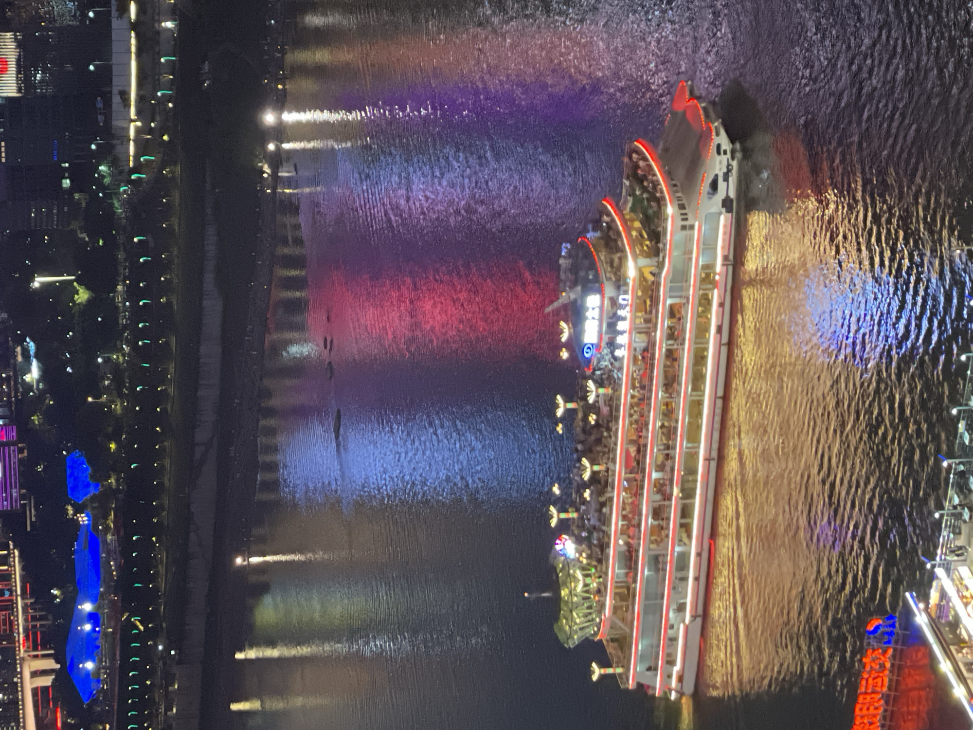 Captured from an elevated perspective at night, the image showcases a river scene in the Yuzhong District, China. A multi-deck passenger ferry, brightly illuminated with red accents and interior lighting that reveals passengers, is traveling on the river. The water reflects a spectrum of colors - red, purple, and blue - likely from the surrounding city lights. The cityscape on the left side of the image features a variety of buildings, some distinctly lit in blue, and streetlights lining the riverbank. The overall composition suggests an active, urban environment.