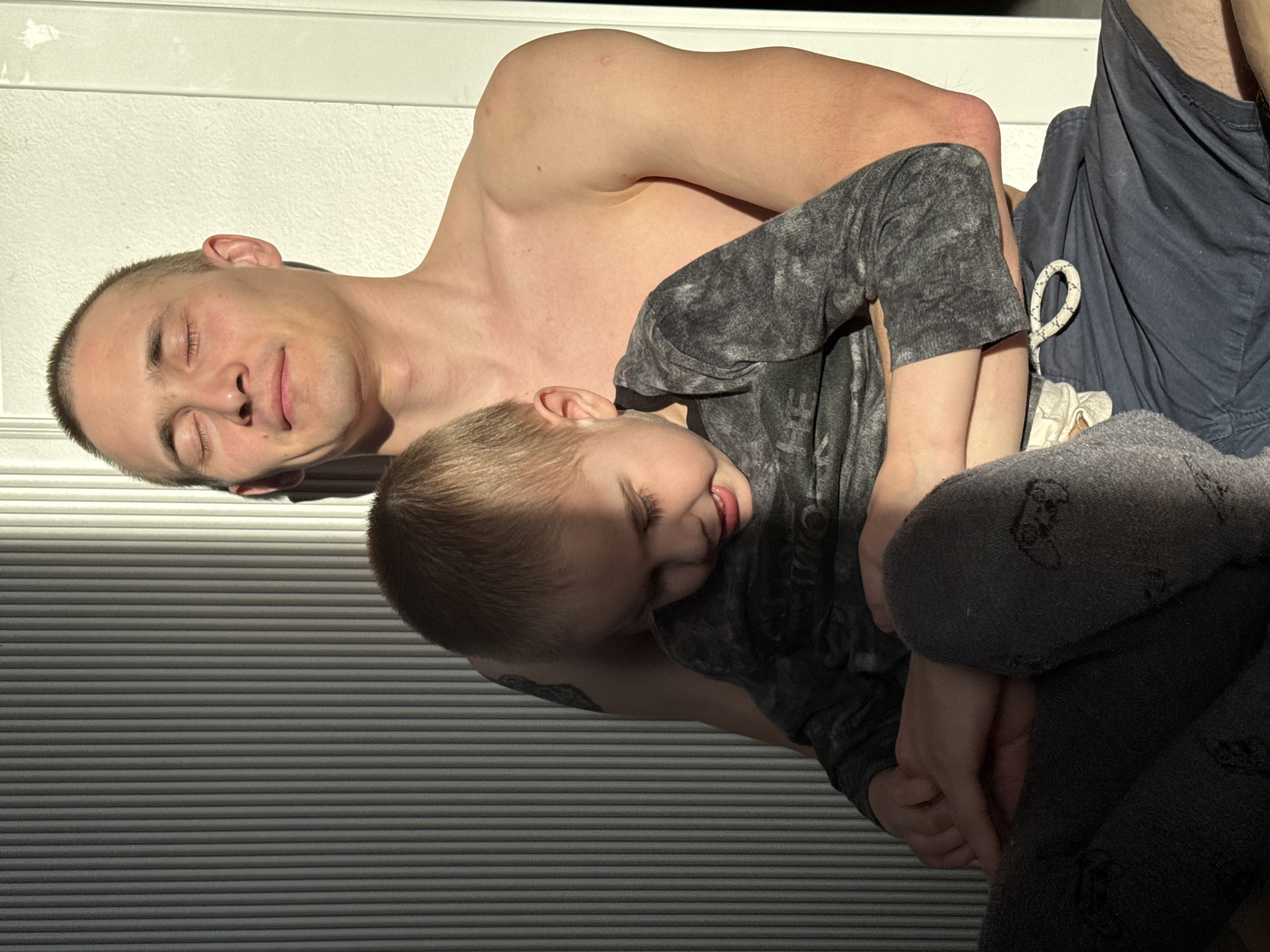 A shirtless man with closely cropped hair is lying down with his eyes closed, holding a young child wearing a dark patterned top and grey leggings. The setting appears to be indoors near a window with closed blinds, casting shadows across the scene. A portion of a person wearing navy shorts is visible to the right. The location is not discernible as Chaikovsky, Russia without additional visual context.