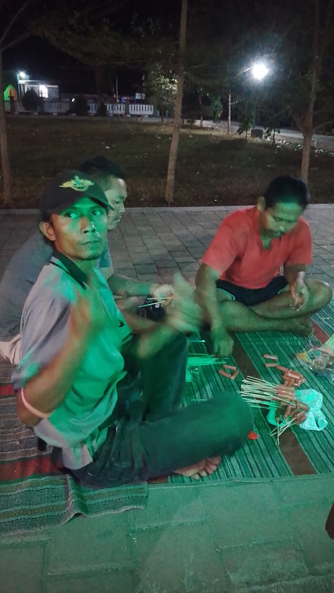 At night in Karanglangu, Indonesia, three men are gathered on a woven mat on a paved area, possibly near a park or public space. The man in the foreground, wearing a black baseball cap and a light-colored polo shirt, is looking directly at the camera. His hands are blurred, suggesting movement, and he is holding a small object. Beside him, to his right, sits a man in a red t-shirt and dark shorts, also looking down and appearing to be working with his hands. Behind them, another man is partially visible, wearing a dark shirt. In front of the men on the mat are several small, uncooked sausages on skewers, as well as some skewers that have already been assembled with sausages. The lighting in the scene is dim, with some artificial lights visible in the background, casting shadows and contributing to the overall green tint of the image. The environment appears to be outdoors, with trees and some buildings visible in the distance.