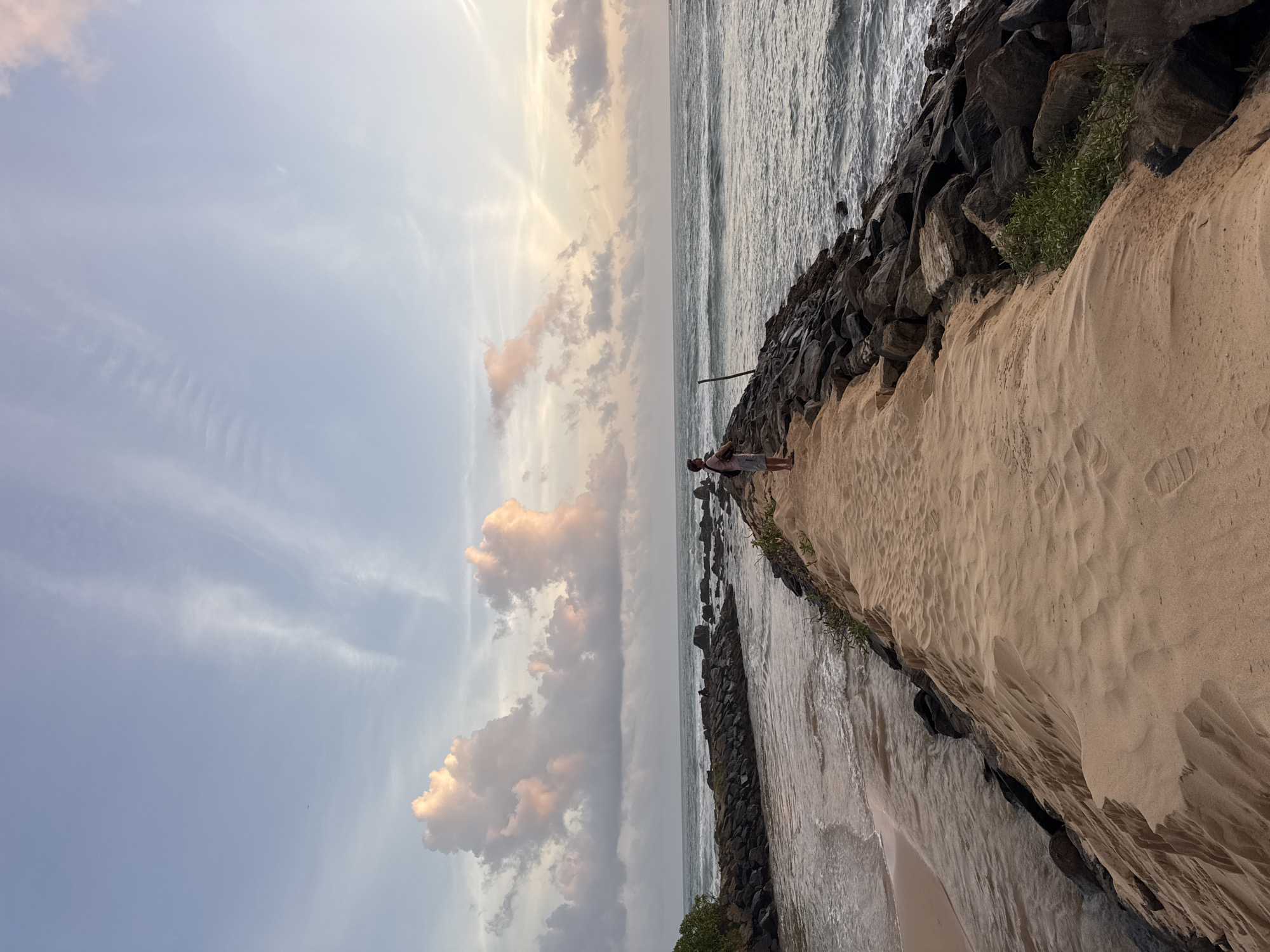 A person stands on a sandy path alongside a rocky breakwater. The path is bordered by dark, rugged rocks on one side and the ocean on the other. The ocean displays gentle waves and the water is a muted gray, reflecting the overcast sky. The sky is a pale blue with streaks of wispy clouds and larger, fluffy clouds tinged with pink and orange, suggesting it is either sunrise or sunset. The overall mood is tranquil and serene. The location is identified as Midigama, Sri Lanka.
