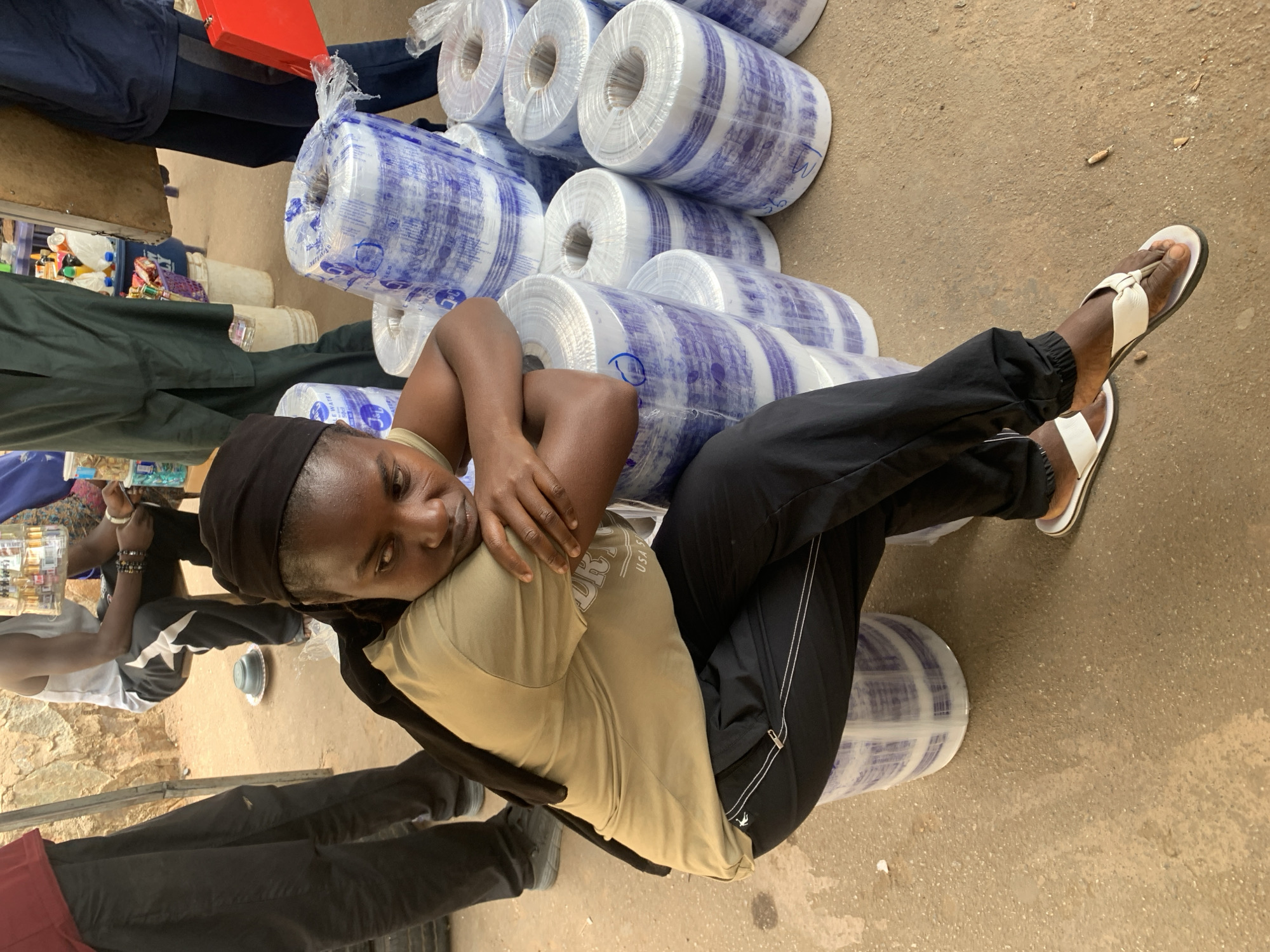 An eye-level shot depicts a person lying on a stack of plastic-wrapped rolls on a concrete surface, presumably in Bukuru, Nigeria. The person has dark skin and wears a black head covering, a tan t-shirt, and black track pants with white stripes. The person's arms are crossed over their chest. The person wears white sandals with a thong strap. The stack of rolls is wrapped in clear plastic with a blue and white pattern. Several people are visible in the background. The floor is concrete. Other items such as boxes, bottles, food, and bags are visible in the background.