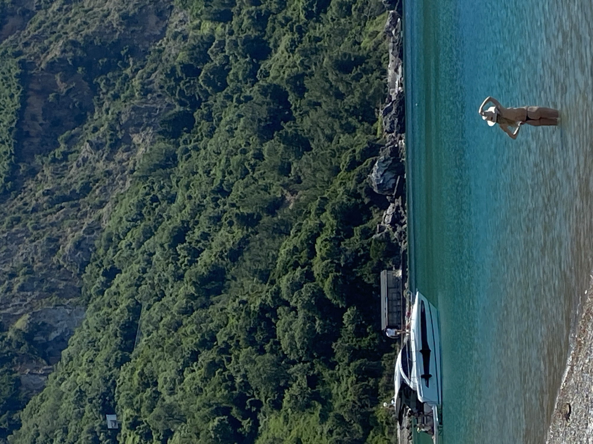 The image depicts a woman wearing a hat standing in the shallow, turquoise water adjacent to a pebble beach in the city of Prijevor, Montenegro. Behind her is a rugged coastline covered in dense green vegetation. Several white speedboats are docked near the shoreline, with a man standing on the dock.