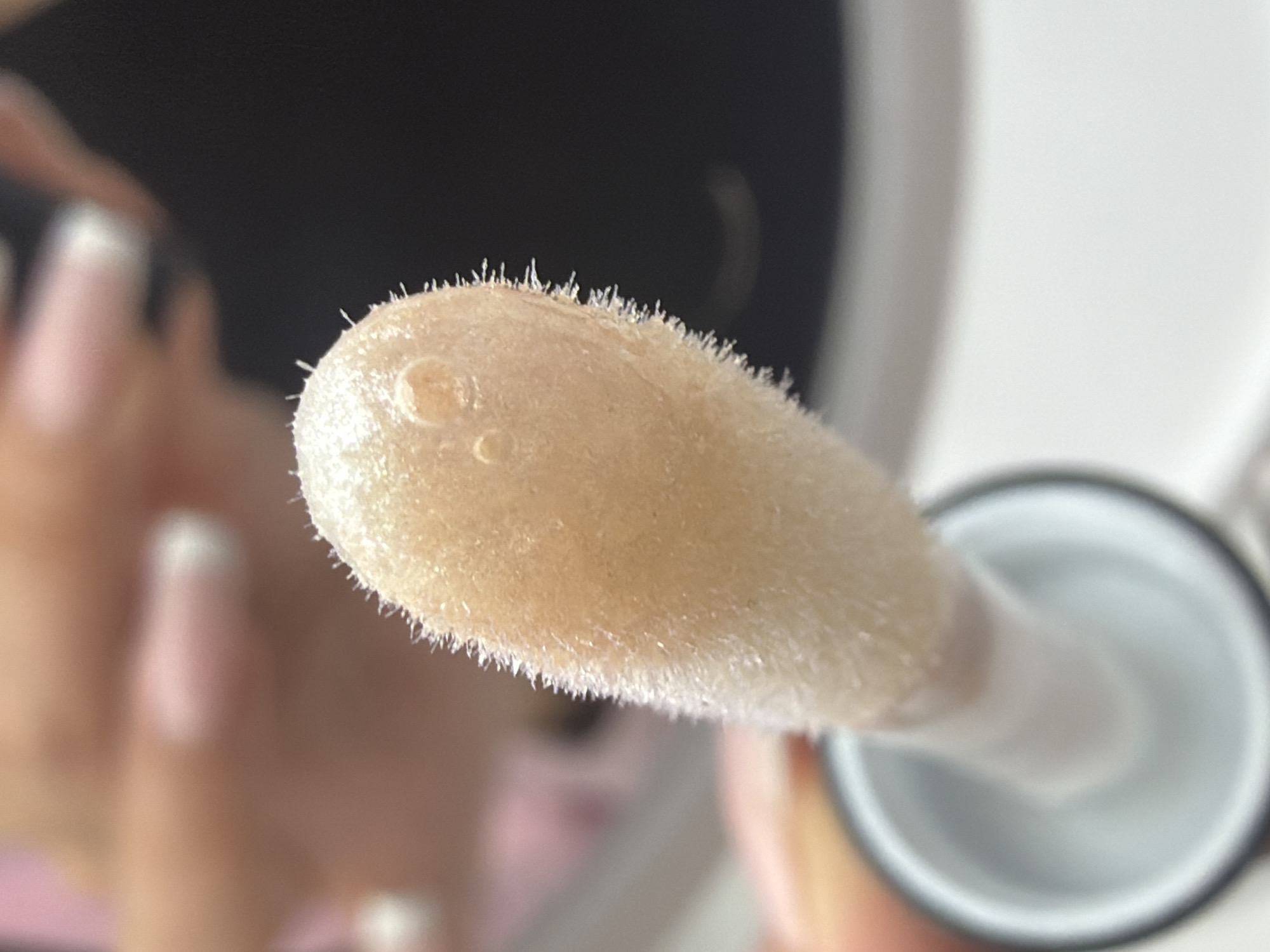 Close-up of a lip gloss applicator covered in product. Two small air bubbles are visible in the product on the applicator. The applicator is being held, and fingers with manicured nails are partially visible. The background is blurry, showing a dark area and a white object. The exact location is unknown.