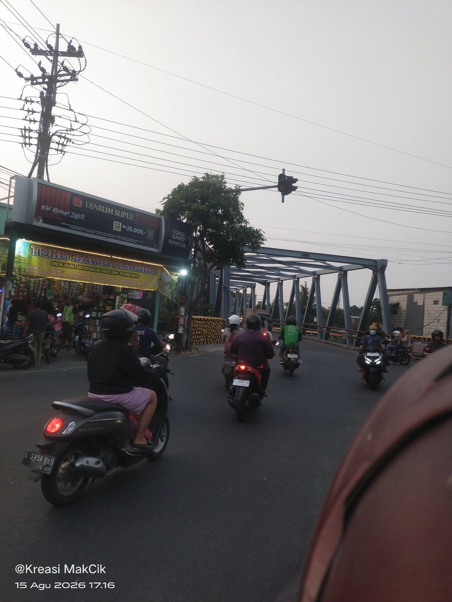 The image depicts a street scene in Wonocolo, Indonesia, on August 15, 2026, at 17:16. The foreground shows the back of a red motorcycle with a rider wearing a dark helmet and light-colored shorts. A black scooter is visible to the left, also with a rider. Several other motorcycles are on the road, with riders wearing helmets, some in green jackets, and others in darker clothing. The road appears to be a main thoroughfare, with a traffic light visible in the distance and a bridge structure overhead. On the left side of the road, there are shops, including one with a prominent sign for "TOKO DAVID RANGGA" and another advertising "DJARUM SUPER." Electrical wires and a utility pole are also present, typical of urban infrastructure. The sky is overcast, suggesting a late afternoon or early evening setting. The overall impression is one of a busy street with moderate traffic.
