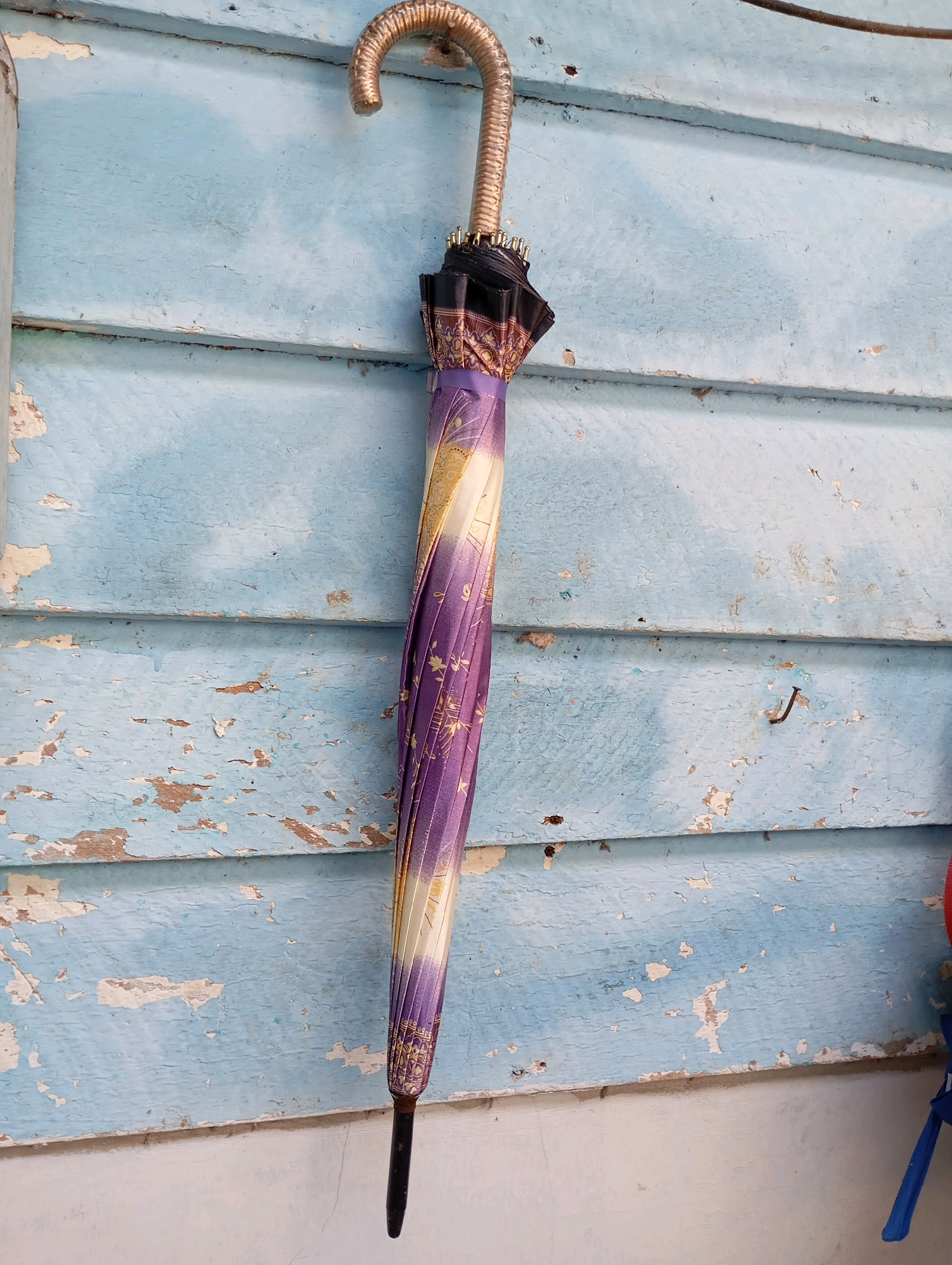A closed, decorative umbrella with a golden handle and a purple and gold patterned fabric hangs vertically against a weathered blue wooden wall. The wall is constructed from horizontal planks, some of which show peeling paint and exposed wood. The umbrella is positioned slightly to the left of the center of the frame. The handle curves over the top plank of the wall. The bottom tip of the umbrella is dark and tapers to a point. The environment appears to be outdoors, with natural light casting subtle shadows on the wall. Part of a white surface is visible at the bottom of the image. In the upper right, a hint of a dark cable is visible. On the far right, glimpses of blue and red fabric suggest other objects are nearby. The scene is located in Balikpapan, Indonesia.