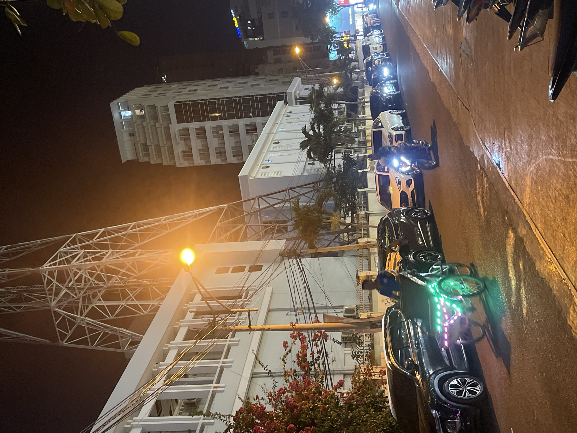 The image captures a vibrant night scene on a city street, likely in Nha Trang, Vietnam. Tall, multi-story white buildings line the left side of the street, with a prominent modern structure featuring multiple windows and balconies. Further down, another taller building with a white facade is visible, along with other urban structures. A large, intricate white metal lattice tower-like structure stands in the foreground on the far left.
The paved street, appearing wet and reflective from the lighting, stretches into the distance on the right. It is lined with parked vehicles, including numerous motorbikes and several cars, some with their headlights illuminated. A white car and a dark-colored sedan are clearly visible in the mid-ground. Next to the dark sedan, a person wearing a blue shirt stands near a bicycle, which is strikingly adorned with bright green and purple LED lights.
The scene is illuminated by numerous streetlights that emit a warm orange glow, casting reflections on the wet pavement and highlighting the architectural details of the buildings. Tropical foliage, including palm trees and bushes with vibrant red flowers, is interspersed along the roadside and between the buildings. The overall ambiance is that of a lively urban environment after dark.
Visible text includes a partially obscured sign on a building in the background that appears to show "GOLDEN" and "COAST," and a blue sign with the letter "H."