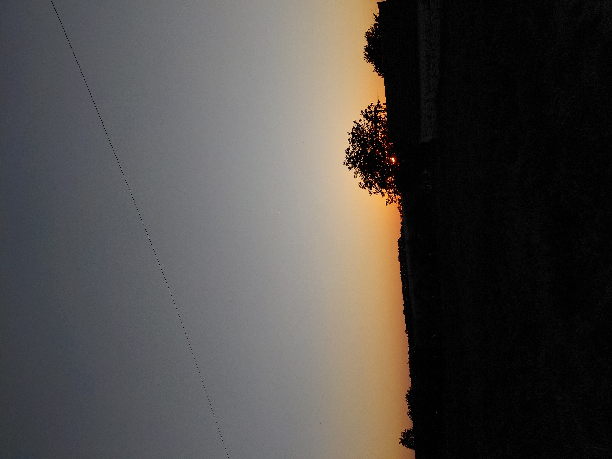 A silhouette of trees and a building is visible against a gradient sky of dusk. The sky transitions from a pale grey at the top to a warm orange near the horizon. A thin wire stretches diagonally across the upper portion of the image. The foreground is completely dark, obscuring any details of the ground. The scene appears to be a rural landscape.