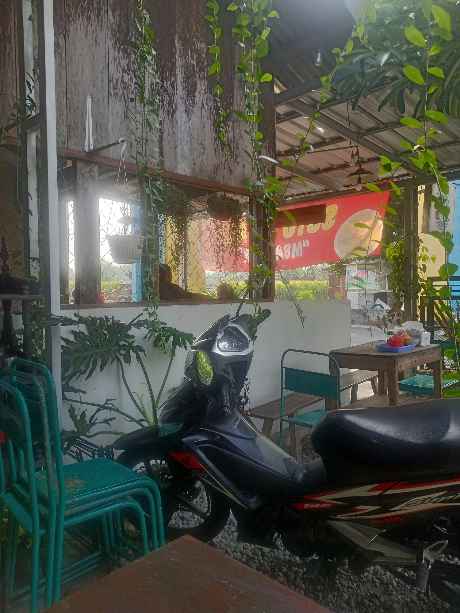 This is a photograph of an outdoor seating area at what appears to be a cafe or eatery in the city of Ngandul, Indonesia. The scene is rustic, with weathered wooden walls and a corrugated metal roof supported by wooden beams. Lush green vines hang from the ceiling and the walls, adding a natural element to the space.
In the foreground, a black motorcycle is parked on a gravel surface. A stack of teal metal chairs is positioned to the left of the motorcycle. A wooden table with a bench and two chairs is set up further back. On the table, there are various condiments in small containers, including red and yellow bowls, and a white object that could be a tissue box or a small appliance.
Through an open window or doorway, a glimpse of the outside can be seen. A red banner with yellow text is hung on the back wall, partially obscured by hanging plants. The overall impression is one of a relaxed, casual outdoor dining environment.