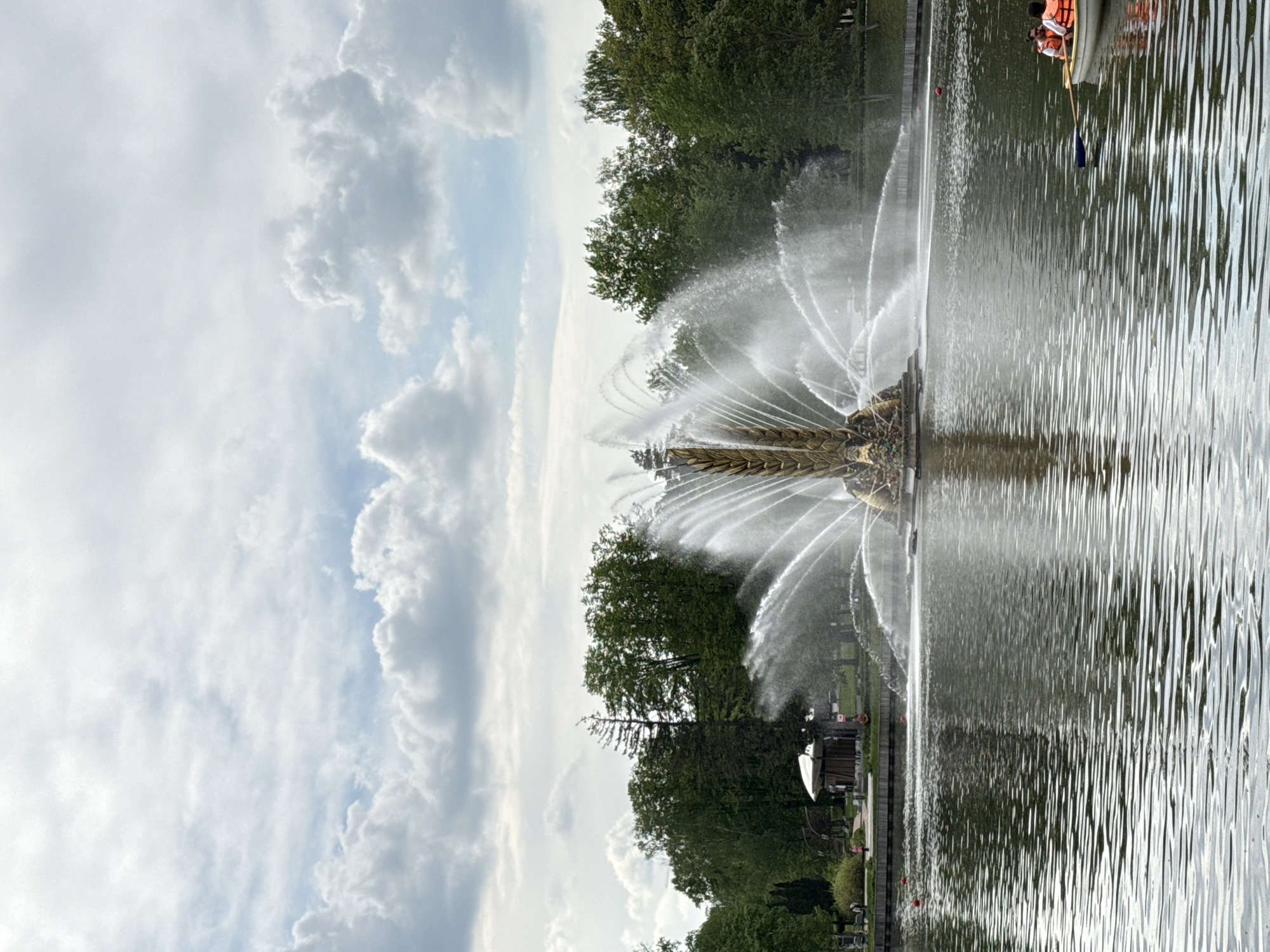 The image shows a fountain in a body of water, possibly a lake or large pond. The fountain structure is elaborate, with water jets spraying outwards in a decorative pattern. Trees and greenery are visible along the edge of the water, suggesting a park or landscaped area. The sky is partly cloudy. The body of water displays linear ripple patterns, and a small boat with passengers wearing life jackets is visible in the background. This is the Golden Ear fountain in VDNKh park, located in Moscow, Russia.