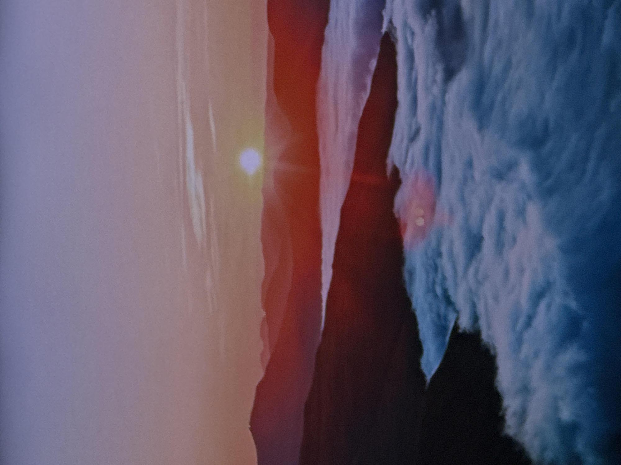 The image depicts a serene mountainous landscape at sunrise. In the foreground, a thick layer of white and blue clouds blankets the lower half of the frame, appearing as a soft, undulating sea. Above the clouds, jagged mountain ranges emerge, silhouetted in deep red and dark purple hues against the vibrant sky. The sun, a bright white orb, is visible just above the horizon, casting a warm, reddish glow over the peaks and illuminating the sky with shades of pink, orange, and lavender. The atmosphere is clear, with a hint of atmospheric haze softening the distant mountains. The scene is devoid of any human presence or man-made structures, focusing solely on the natural grandeur of the mountains and the dawn sky. The image does not contain any elements that specifically place it in the city of Sinyavino, Russia.