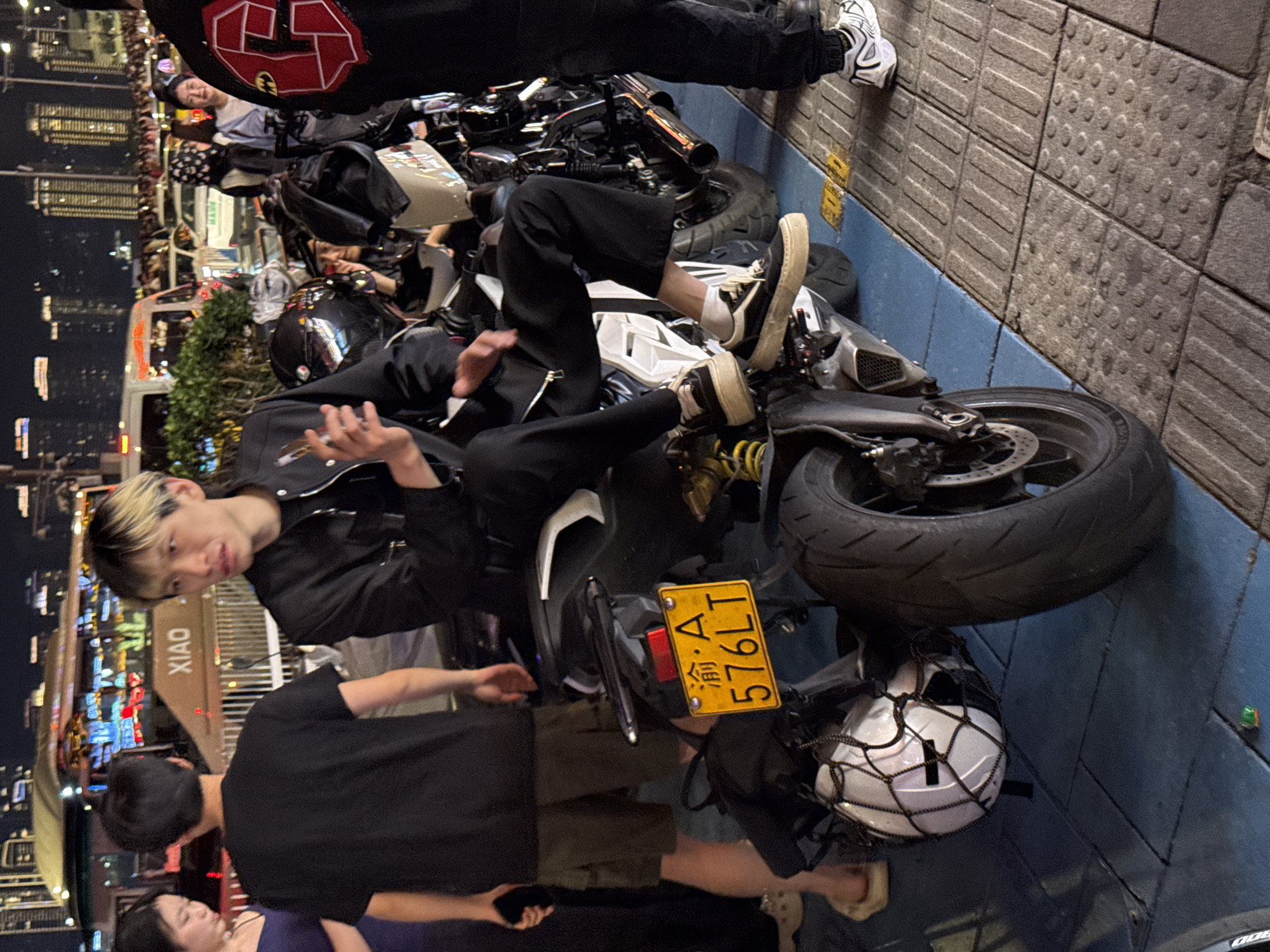 The image depicts a group of people gathered around parked motorcycles in Yuzhong District, China. One individual with blonde-streaked hair is sitting on a motorcycle, wearing a black jacket and dark pants. Another person in a black t-shirt and khaki shorts stands nearby, holding a phone. A motorcycle in the foreground has a license plate reading "渝A·576LT." Several other motorcycles are visible in the background, and a helmet rests on the ground near the first motorcycle. In the background, city lights and illuminated buildings suggest an urban setting.