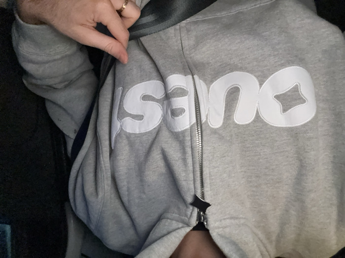 An individual is depicted from the upper torso, seated within a vehicle in Givors, France. The person wears a light gray zippered hoodie featuring white, puffed-style lettering on the front, spelling "DUES." The 'D' in "DUES" has a star-shaped cutout. The hoodie's zipper pull is a black four-pointed star. A dark-colored seatbelt crosses diagonally from the individual's left shoulder to their right hip. The individual's right hand, adorned with a thin gold-colored ring on the ring finger, is holding the lower strap of the seatbelt. The background is dark and indistinct, consistent with a vehicle interior.