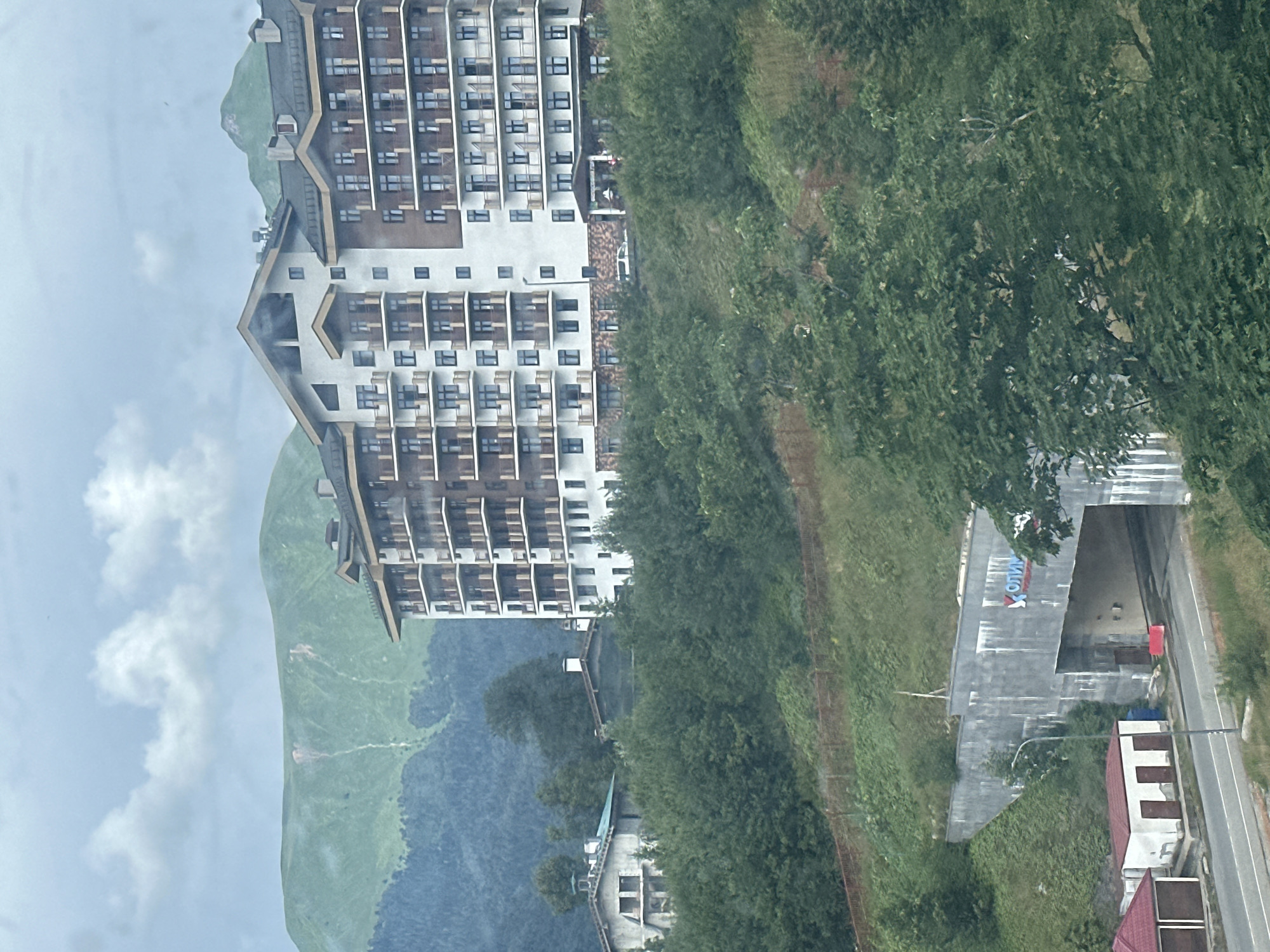 The image captures a scenic view of the resort town of Estosadok, Russia, nestled amidst lush green mountains. In the foreground, a modern, multi-story apartment building with balconies stands prominently on a hillside, its architecture blending with the natural landscape. Below it, a tunnel entrance is visible, marked with the stylized "Y" logo of Olympstroy, the company responsible for constructing Olympic venues in Sochi. The tunnel is flanked by a road and a few small buildings with red roofs. The background is dominated by rolling, verdant mountains, partially obscured by clouds, suggesting a potentially overcast or misty day. The overall impression is one of a well-developed mountain resort area, integrating modern infrastructure with natural beauty.