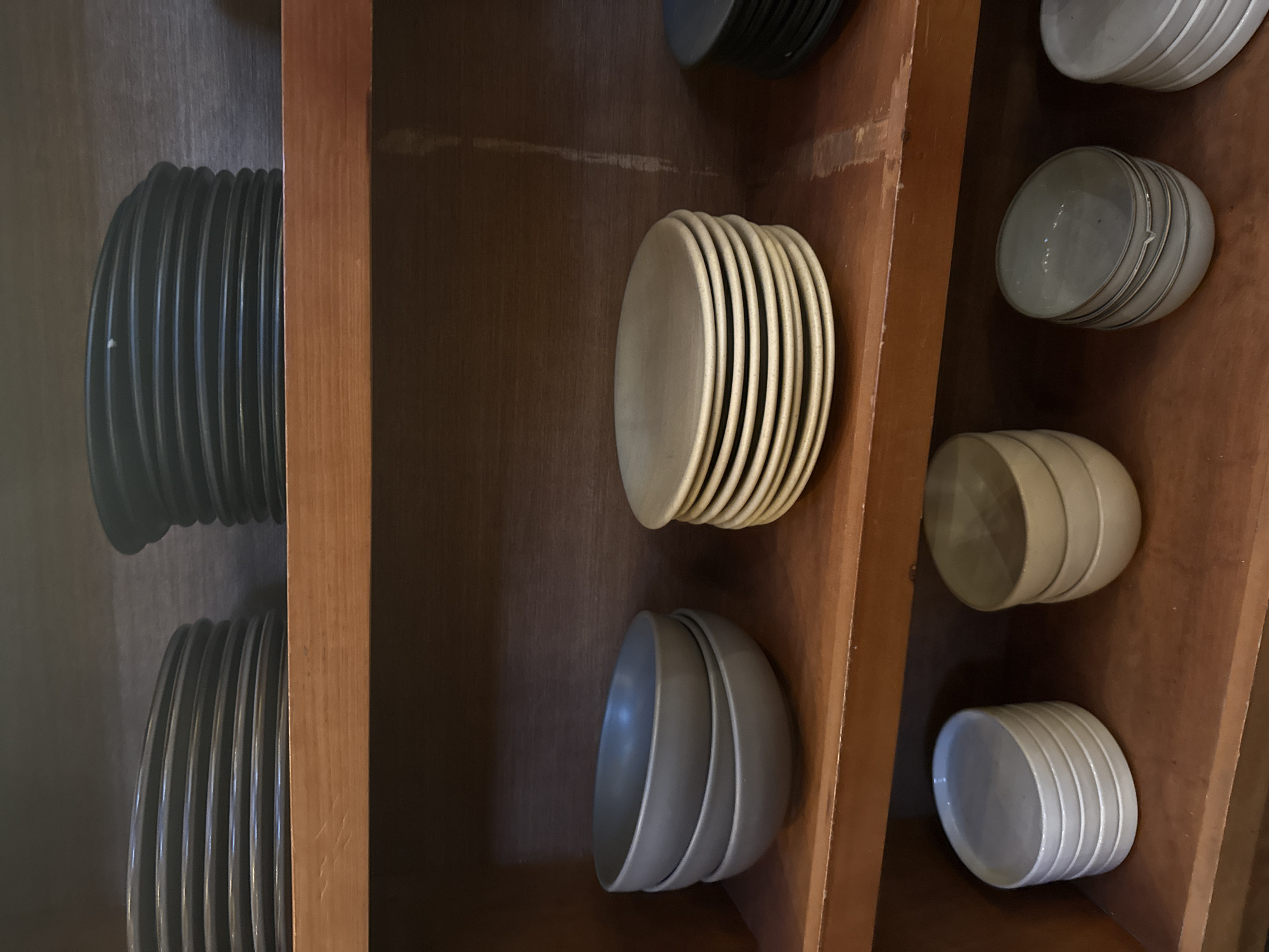 The image shows a wooden shelf containing stacks of plates and bowls. On the left, there are stacks of dark-colored plates. In the center, there is a stack of light-colored plates and a stack of bowls. On the right, there are stacks of bowls and plates in shades of white or light gray. The shelf appears to be part of a built-in structure.
There is no information about the location in the city of San Cristóbal, Mexico, or any information about people, subjects, objects, actions, interactions, and the environment to provide a concise, forensic, matter-of-fact description of the scene.