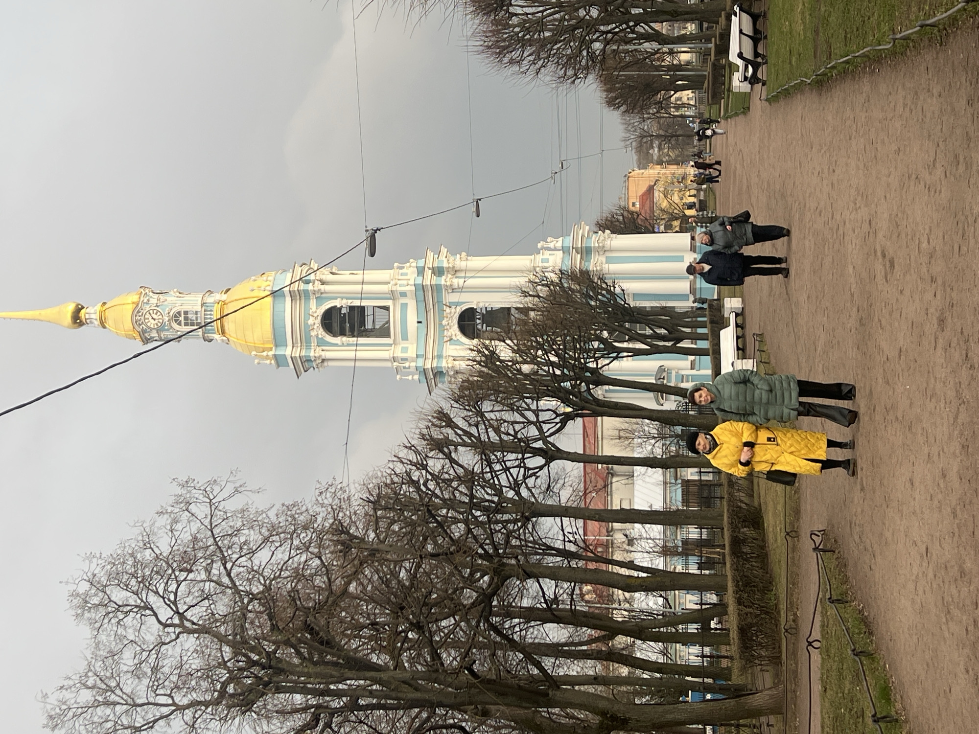 The image depicts a park in Saint Petersburg, Russia, featuring the Naval Cathedral of Saint Nicholas in the background. The cathedral's golden dome and blue-and-white facade are prominent. In the foreground, a gravel path is lined with bare trees and benches. Groups of people are walking along the path, including a group of three standing closer to the camera. One individual is wearing a yellow coat, while another wears a green coat, and the third is wearing a dark coat. Power lines are visible overhead. The sky is overcast, suggesting a cloudy day.