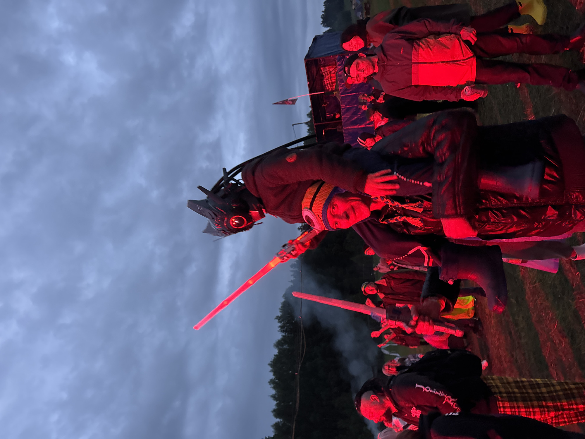 The image captures an outdoor night gathering in a grassy field in Большая Кивара, Russia, illuminated by a dominant red artificial light source. In the foreground, a man with a multicolored beanie (blue, yellow, orange) and a dark jacket carries another individual on his shoulders. The person being carried wears a dark hooded jacket and a prominent black, futuristic helmet with a single glowing red eye and various external components. This individual holds a long, glowing red prop energy sword upright in their right hand and a shorter, similar glowing red prop weapon pointed downwards in their left hand.
To the left of the central figures, a man with dreadlocks, a beard, a dark hoodie bearing the text "Jollity," and plaid pants is partially visible. Further into the mid-ground, numerous individuals are present, some with raised arms, all bathed in the red illumination. A man in a two-tone jacket (dark top, orange/red bottom) and a baseball cap holds a can on the right side of the mid-ground. On the far right, a woman in a dark jacket and bright yellow rubber boots is visible. Smoke, emanating from an unseen source, rises on the left side of the frame, illuminated by the red light. In the background, a makeshift stage or structure, constructed with blue tarps and a tent-like roof, is visible to the right. A flag on a pole stands near this structure. A dense line of trees forms the distant horizon under an overcast, dark sky.