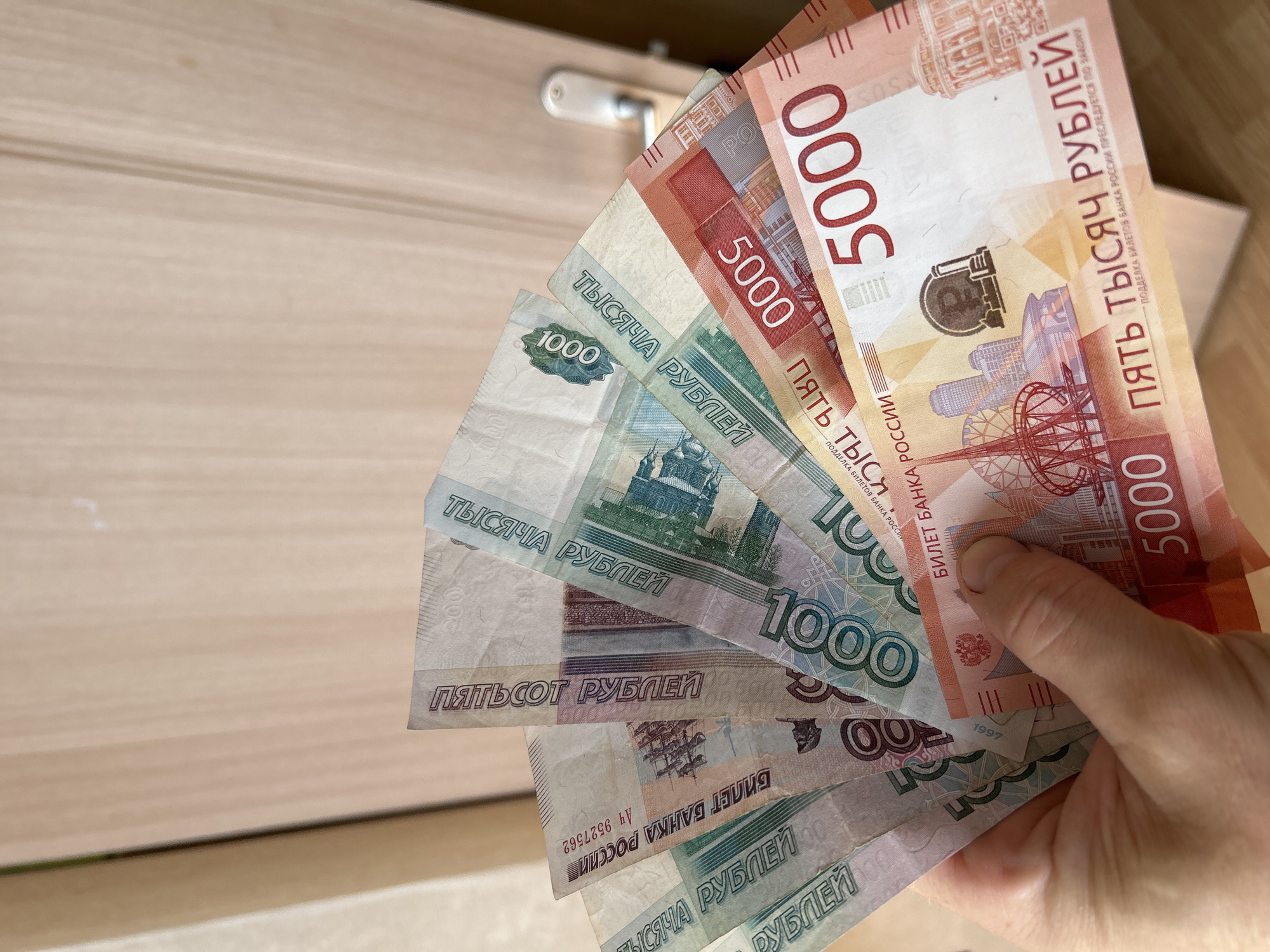 The image depicts a hand holding a fan of Russian Ruble banknotes. The denominations visible include 500 Ruble, 1000 Ruble, and 5000 Ruble notes. The 5000 Ruble notes show landmarks from the city of Khabarovsk. The 1000 Ruble notes show a depiction of Yaroslavl, Russia. The background appears to be a light-colored wooden surface with a handle.