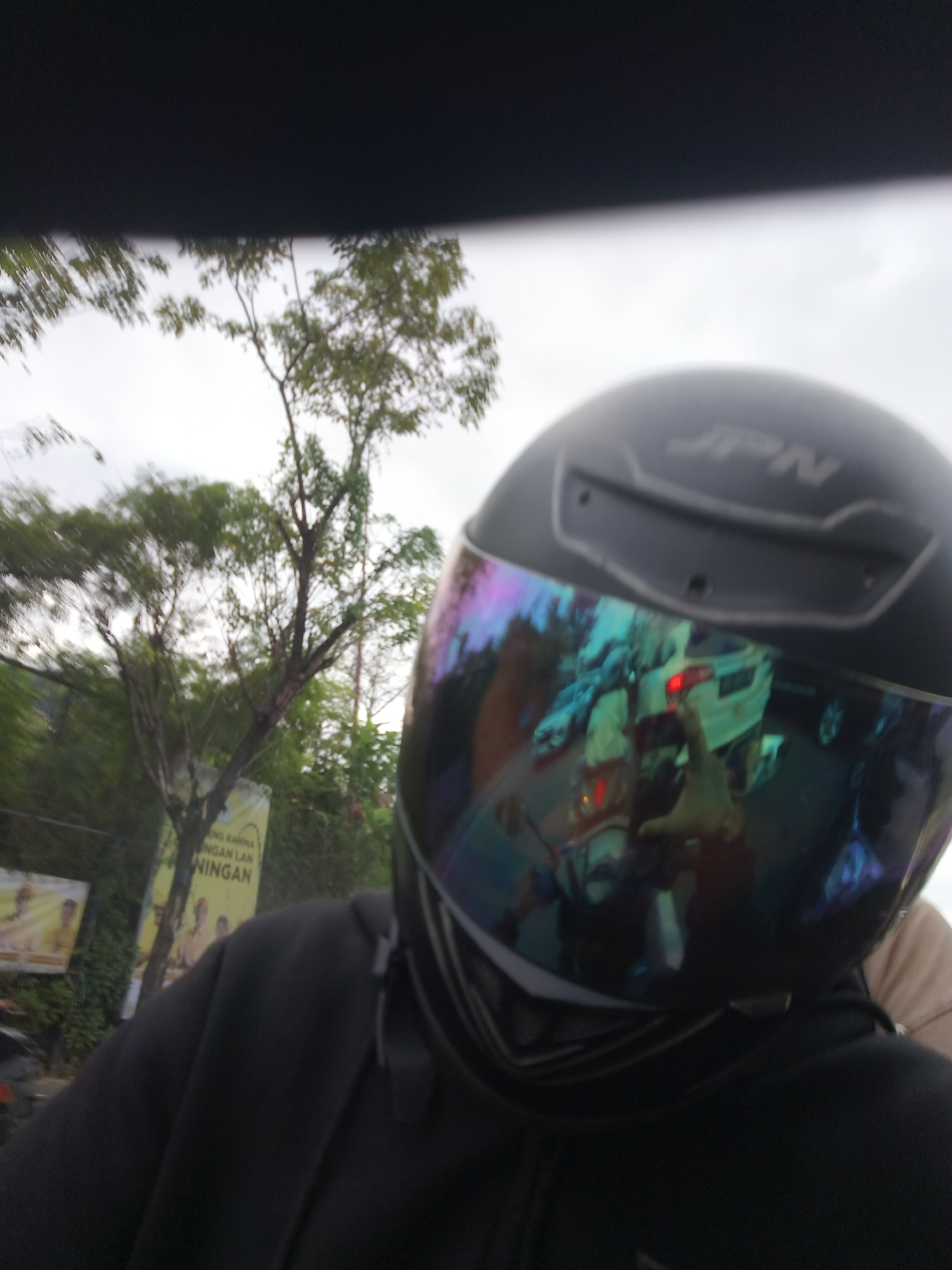 The media file is a first-person perspective photo taken from the viewpoint of someone wearing a black motorcycle helmet with a reflective, iridescent visor. The helmet has the "JPN" logo visible on its surface. The wearer is wearing a black hoodie.
The scene appears to be outdoors, with trees and foliage visible in the background. The sky is overcast, suggesting a cloudy day. In the reflection of the visor, several objects are visible, including a white vehicle with its brake lights illuminated, other vehicles, and what appears to be a hand reaching towards the camera. There are also two posters visible in the background. One poster is yellow with text, and the other is a portrait-style advertisement.
The context provided indicates the location is Kuta Selatan, Indonesia. The overall impression is that the photo was taken while riding a motorcycle on a street, possibly in an urban or suburban area, under cloudy weather conditions. No specific emotions are discernible from the image.
Visible text includes "JPN" on the helmet and some Indonesian text on the yellow poster, which is partially obscured and illegible in the provided image.