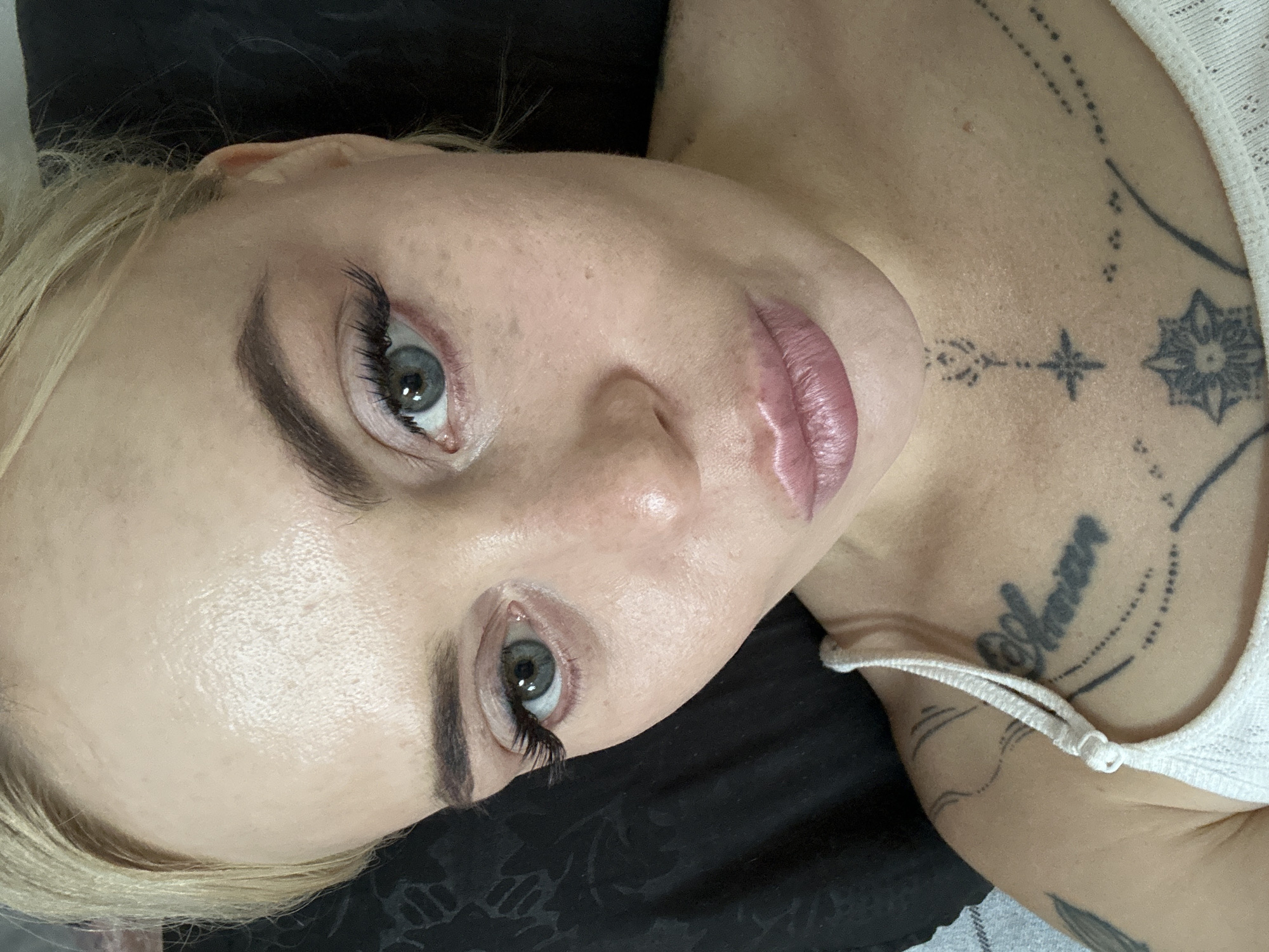 A close-up, high-angle, over-the-shoulder selfie is captured of a fair-skinned Caucasian woman with blonde hair pulled back, her face tilted slightly upwards and to her right. She is lying down against a dark background, possibly a pillow or blanket, with a sliver of a gray, checkered pattern visible in the lower left corner, indicating an indoor setting. Her eyes, with blue-green irises, are wide open and looking towards the camera, framed by dramatic false eyelashes and well-defined eyebrows. Her lips are slightly parted, revealing a subtle pout, and are adorned with a shimmery pink lipstick. Her skin exhibits a light dusting of freckles across her nose and cheeks, and appears dewy, with subtle highlights on her forehead and cheekbones. She is wearing a white tank top, the strap of which is visible over her left shoulder. Several tattoos are visible on her upper chest and collarbone area, including dark ink lettering and geometric designs. The overall lighting is soft and diffused, casting gentle shadows and highlighting the contours of her face. The image is in focus, with a shallow depth of field that blurs the background.