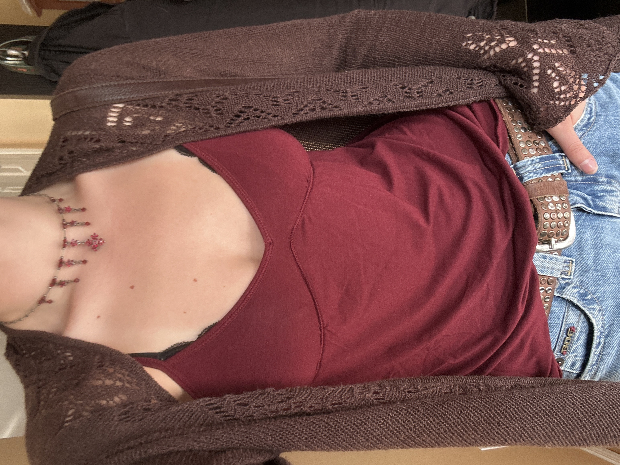 A close-up, angled shot captures the torso of a person, likely reclining or lying down, dressed in a casual outfit indoors. The person's face is not visible, focusing on their clothing and accessories from the chest down to the upper thighs.
The individual wears a dark red or maroon camisole or tank top, featuring a subtle V-neckline. A hint of black lace trim is visible near the bra line. Over this, a dark brown, open-knit cardigan or shrug is draped, featuring intricate lace-like patterns along the sleeves and trim.
Below the red top, light blue denim jeans are visible, along with a prominent brown leather belt adorned with numerous silver studs. A person's hand with unpolished fingernails rests near the belt and jeans.
A silver-toned necklace with multiple red beads or charms hangs around the person's neck, extending onto the upper chest. The person's fair skin has a few small moles visible.
In one of the cropped images, the text "BDG" is visible on the pocket of the jeans. The overall lighting is soft and even, suggesting an indoor setting without specific time-of-day indicators. No specific location cues pointing to Kelowna are visible within the image itself.