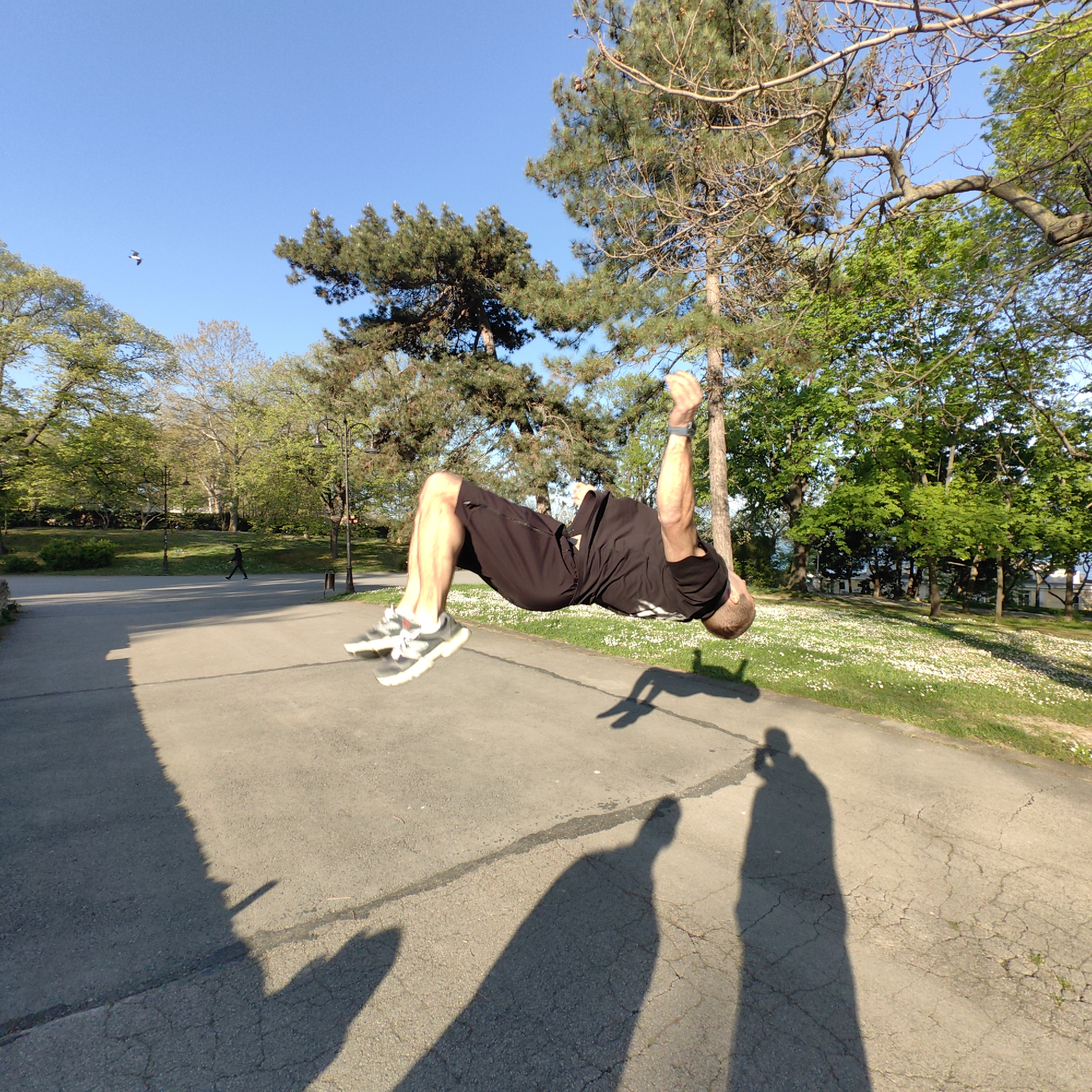 A man is performing a backflip on an asphalt path in a park. He is wearing a black t-shirt, black shorts, and gray athletic shoes with white soles. The shadow of the man and two other figures are visible on the ground. The park features trees, a patch of white flowers, and a person walking in the background. The sky is blue with a few clouds. The scene is taking place in Varna, Bulgaria.