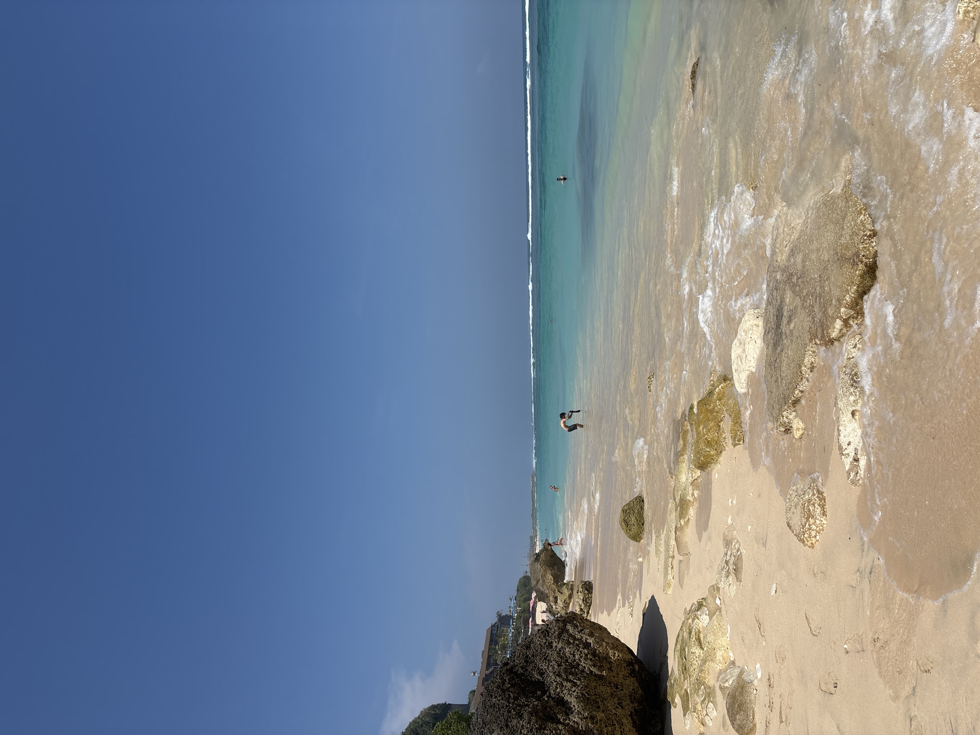 This image depicts a sunny beach scene in Ungasan, Indonesia. The sky, occupying the upper half of the frame, is clear and uniformly blue. The ocean water transitions from a clear, light turquoise near the shore to a deeper blue further out, with visible white-capped waves in the distance.
The shoreline consists of light beige sand interspersed with numerous rocks of varying sizes and colors. In the immediate foreground, a large, dark brown boulder is positioned on the sand in the lower left. Numerous other irregular-shaped rocks, predominantly light tan to yellowish-brown, are scattered along the sandy beach and partially submerged in the shallow, clear water. Small, foamy waves are observed breaking and receding over these rocks and the wet sand.
Approximately eight individuals are present. A male figure, wearing dark shorts, stands in knee-deep water in the mid-ground, bending forward at the waist. Another person is visible in the deeper water further out, appearing to be swimming or wading. Several other individuals are scattered along the left side of the beach and in the shallow water, some near a cliff face.
Along the upper portion of the cliff face on the left, a multi-story building with a brown roof and light-colored walls is visible, surrounded by green foliage. Below this building, on the beach, several red beach umbrellas are discernible. The scene is brightly lit by direct sunlight, casting clear shadows from the rocks.