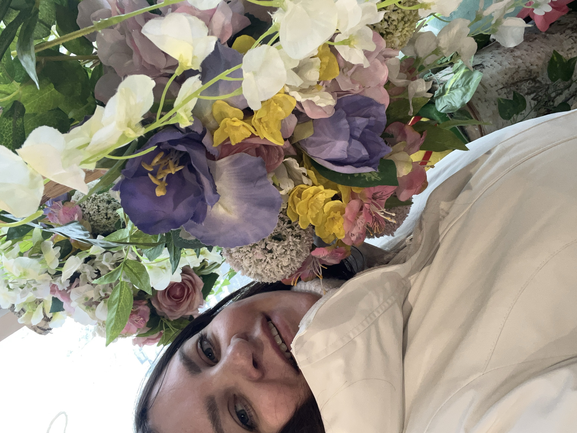 The image displays a brightly lit, portrait-oriented shot featuring a woman smiling beneath a vibrant overhead floral arrangement. The photograph is rotated 90 degrees counter-clockwise, showing the woman's head on the right side of the frame, looking upwards with a joyful expression.
The woman has dark hair and light-colored eyes, and a wide, genuine smile that reveals her teeth. She is wearing a light-colored, possibly white or off-white, collared jacket or top. Her face is illuminated from above, highlighting her cheerful demeanor.
Dominating the upper and central portions of the frame is a dense, multi-colored display of artificial flowers. The arrangement includes a variety of blossoms in white, bright yellow, different shades of purple (such as lavender and a deeper violet), and soft pink. Green leaves and foliage are intricately woven among the flowers, adding to the lush appearance. Closer inspection reveals the synthetic nature of the petals and leaves.
The background behind the flowers is softly out of focus, suggesting an indoor setting or a shaded area. The overall scene conveys a lighthearted and pleasant atmosphere. No text is visible in the image. This moment captures a candid interaction with a decorative floral display, situated in Perm, Russia.