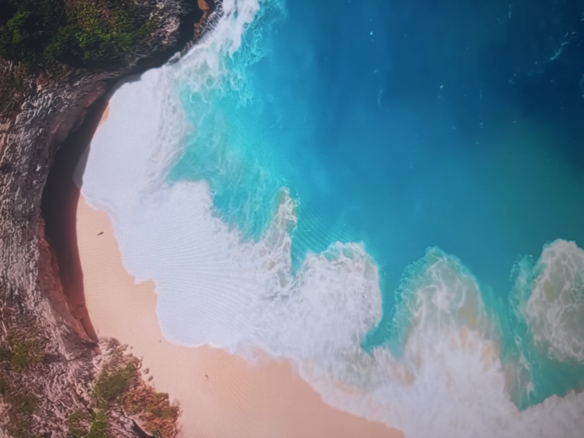This is an aerial, high-angle shot of a tropical beach with turquoise water and white waves. The beach is lined with rocky cliffs covered in lush green vegetation. The sand is a pale peach color. The water transitions from a vibrant turquoise near the shore to a deeper blue further out. There is a small, dark silhouette of a person on the beach. The scene depicts a serene natural landscape. This image is not from Almaty, Kazakhstan; Almaty is a landlocked city in a mountainous region and does not have a tropical beach.