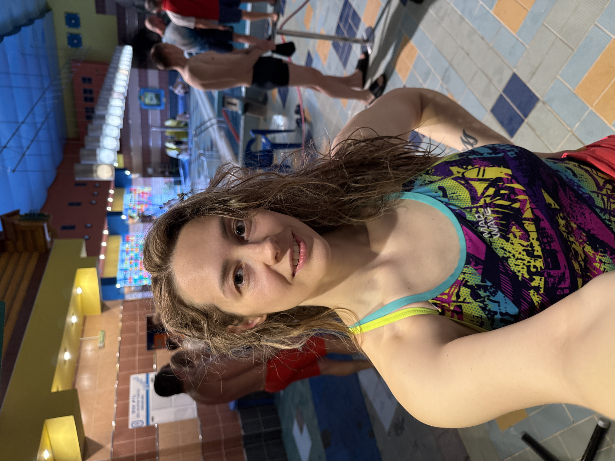 A selfie taken at the water park "Izumrudny" in Chaikovsky, Russia, shows a woman with wet, wavy brown hair and light skin. She wears a brightly patterned swimsuit with blue and yellow straps. The background includes several people in various states of undress near a pool with a brightly colored tiled floor. There are structures reminiscent of cityscapes with multiple windows and colorful panels. The building's architecture suggests a play area within the water park.