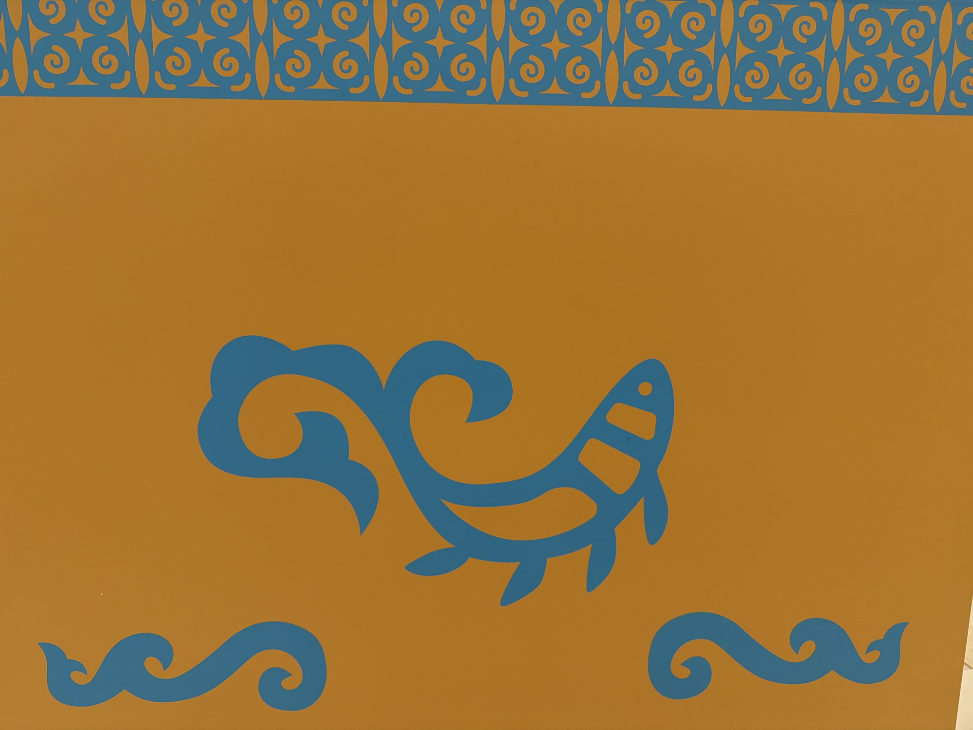 The image is a close-up shot of an orange wall decorated with blue patterns. The top of the wall features a border of geometric shapes and swirls in blue. Below this border, a large stylized blue fish is depicted in the center of the frame. To the bottom left and right of the fish, there are two curved, wave-like blue patterns. The overall aesthetic is decorative and appears to be a mural or painted design. This image was taken in Yuzhno-Sakhalinsk, Russia.