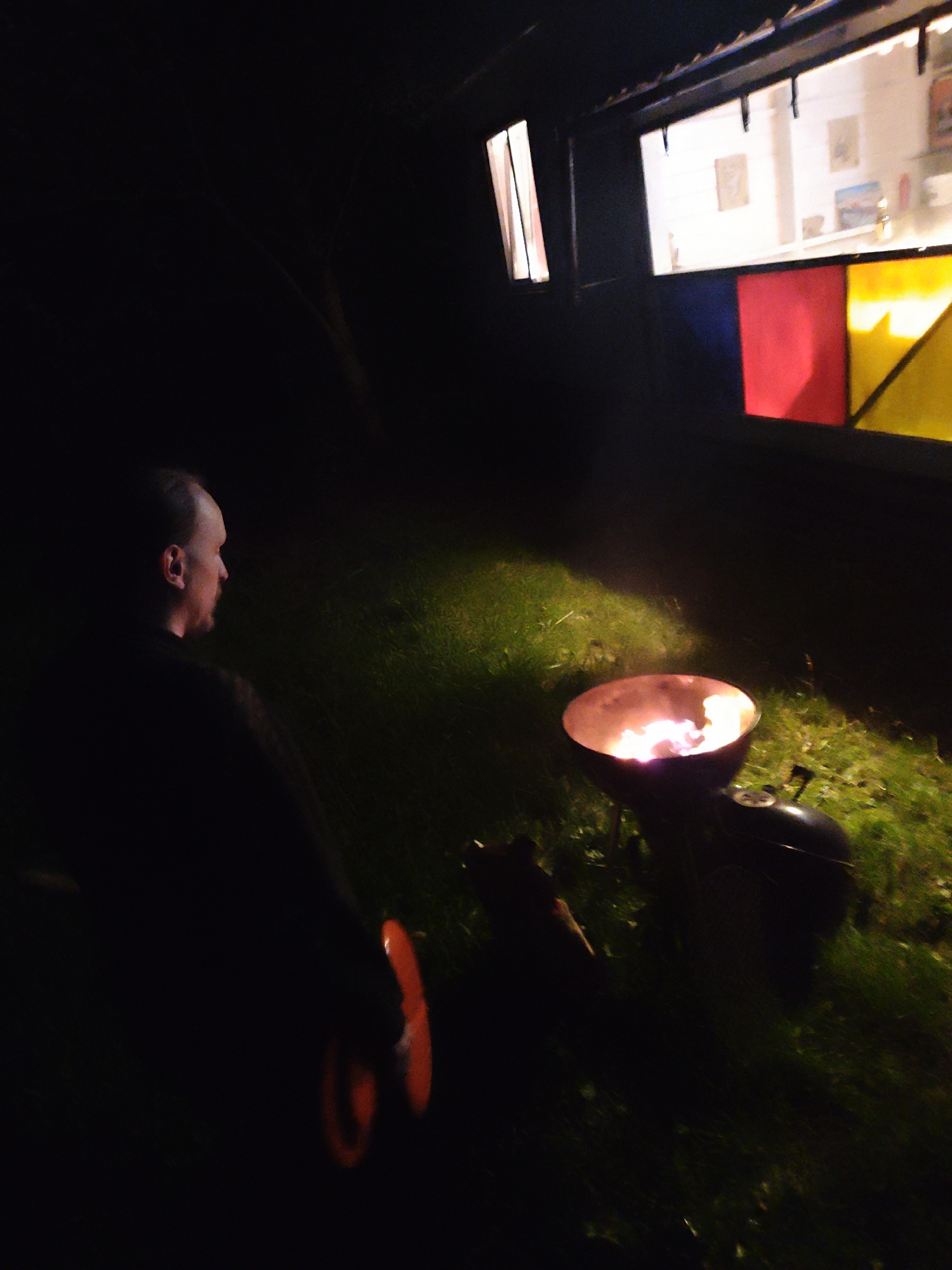 The photograph depicts an individual standing outdoors in the vicinity of Свистуха (кузяевский С/о), Russia, at night. To the left of the frame, a person is partially visible, holding an orange object that appears to be a frisbee or disc. In the foreground, a charcoal grill is actively burning, casting a glow onto the surrounding grass. In the background, there is a structure or building with windows, one of which has stained glass panels in red, yellow, and another indiscernible color. The other windows reveal the interior space to be lighted. The general environment suggests a private property or backyard setting.