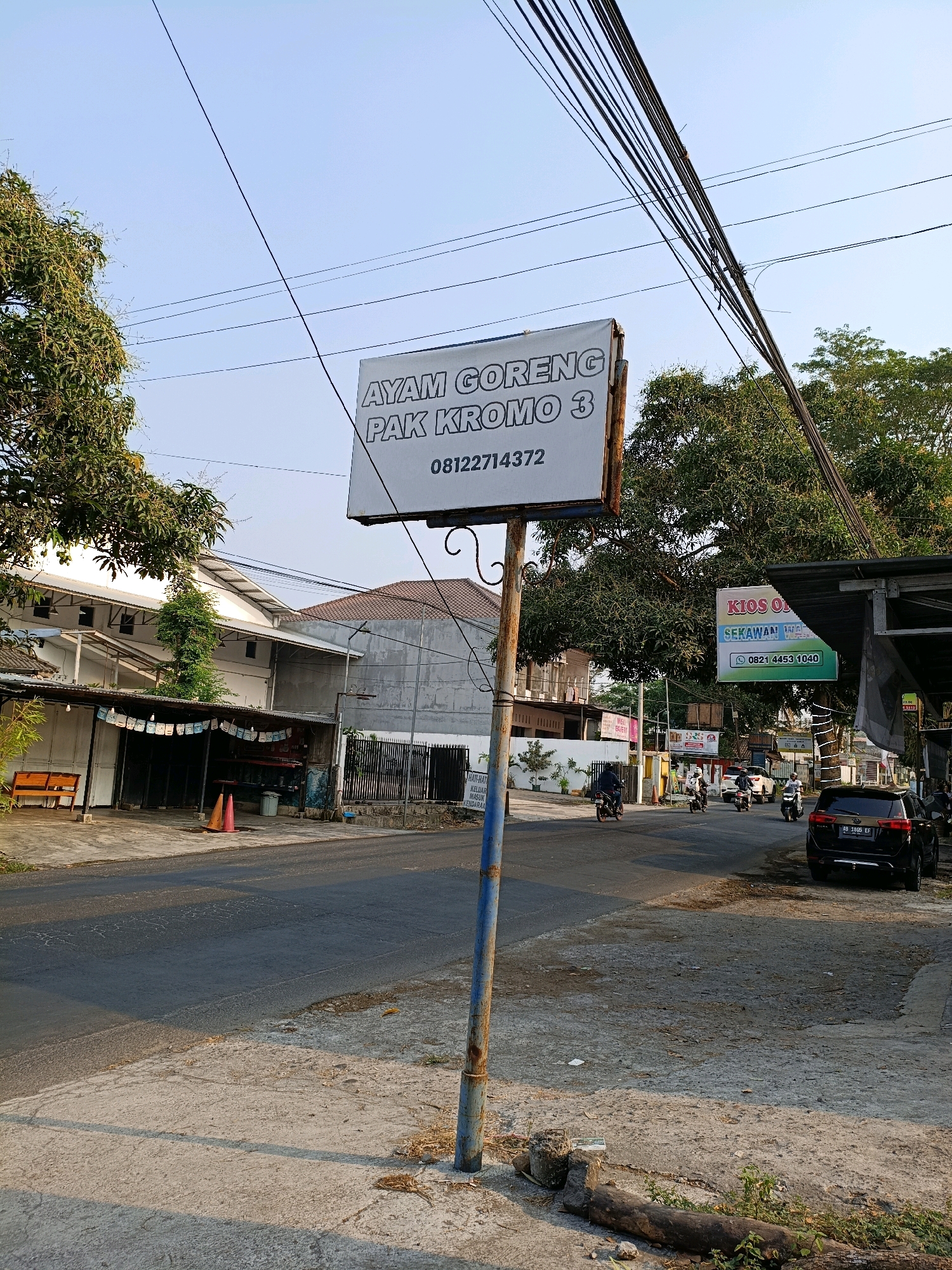 The image depicts a street scene in Ngemplak, Indonesia, captured during daylight hours. In the foreground, a paved road runs horizontally, with a concrete sidewalk to the left and a dirt shoulder to the right. A weathered metal pole supporting a white sign with black text is prominently located on the sidewalk. The sign reads "AYAM GORENG PAK KROMO 3" and below it, the phone number "08122714372". Further down the street, on the right side, another sign is visible, green with white and colored text, advertising "KIOS OK SEKAWAN WA" with the phone number "0821 4453 1040".
A black SUV with license plate "AB 1885 EF" is parked on the right side of the road. Several motorcycles are in motion on the road, some with riders. To the left of the road, there is a building complex with a white roof and open-air structures, suggesting commercial activity. Trees with dense foliage are present on both sides of the street, and some are also visible behind the buildings. Power lines crisscross the sky overhead. The overall environment appears to be a suburban or semi-urban area.
