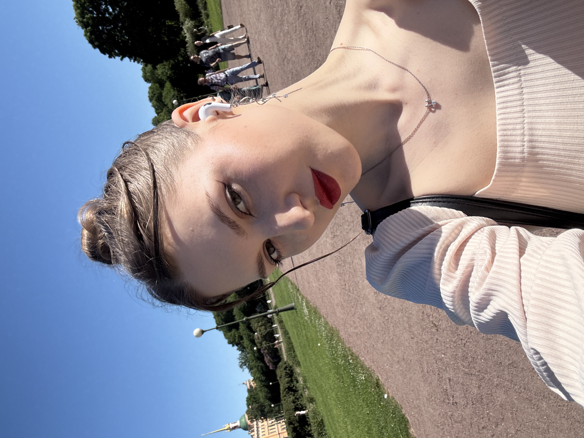 A selfie-style portrait captures a young woman outdoors under a clear blue sky, with the Mikhailovsky Castle (St. Michael's Castle) visible in the background on the far left. The scene is set in Saint Petersburg, Russia, within a park or garden area.
The woman, centrally positioned and slightly angled, looks directly at the camera. She has light brown hair styled in a messy bun, with some loose strands framing her face. Her makeup includes defined eyebrows, dark winged eyeliner, and dark red lipstick. She wears a white wireless earbud in her right ear and a long, silver-toned dangle earring with a spiral design and small crystals on the same side. A delicate silver-colored necklace with a small pendant, featuring two interlocking elements (one with a clear stone), rests on her décolletage. She is dressed in a light pink or peach ribbed off-the-shoulder top. A black strap, possibly from a bag or undergarment, is visible on her left shoulder.
The background features a wide, unpaved light brown pathway that curves to the right, flanked by green grass and lush trees and bushes under bright daylight. A black lamp post with a white spherical light fixture stands to the left of the path. In the far left distance, the distinct yellow-orange walls, green roofs, and slender green spire of the Mikhailovsky Castle are clearly identifiable. Several blurred figures, appearing casually dressed, are walking along the pathway in the mid-ground. Sunlight casts distinct shadows on the woman's clothing and neck.