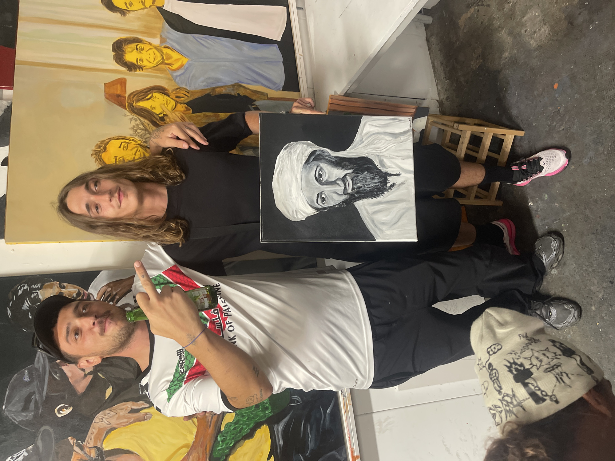 The scene depicts two men in an art studio, located in Marseille, France. The man on the left, estimated to be in his late 20s or early 30s, has a short dark beard and mustache, with short dark hair partially concealed by a black baseball cap worn backward. He wears a white short-sleeved athletic shirt featuring the Palestinian flag design (red triangle, black, white, and green stripes), "Capelli Sport" branding, and Arabic text reading "فلسطين" (Palestine) along with "BANK OF PALESTINE". He is also wearing black athletic shorts and gray chunky athletic shoes. He holds a green glass beer bottle in his left hand, points with his right index finger, and has his left arm draped over the shoulder of the man on the right.
The man on the right, also in his late 20s or early 30s, has long wavy light brown hair and is clean-shaven. He wears a black short-sleeved t-shirt, black shorts, black socks, and pink and white athletic shoes. He holds a small square black-and-white canvas in both hands, presenting it towards the viewer. The painting is a head-and-shoulders portrait of a bearded man wearing a white turban, resembling Osama bin Laden, with a faint signature, possibly "Cyril Albert," in the bottom right corner. His right arm is around the left man's shoulder.
In the background, two large paintings are visible. The canvas behind the left man features abstract figures rendered in dark, yellow, and green tones, with one figure wearing a black baseball cap. The canvas behind the right man displays several stylized human figures in yellow, brown, and light blue against a light tan background, with a lamp-like object in the upper center. The floor is concrete with scattered paint specks. A light-colored beanie adorned with black doodle-style drawings rests on the floor in the bottom left. A small wooden stool and a white table are also present.