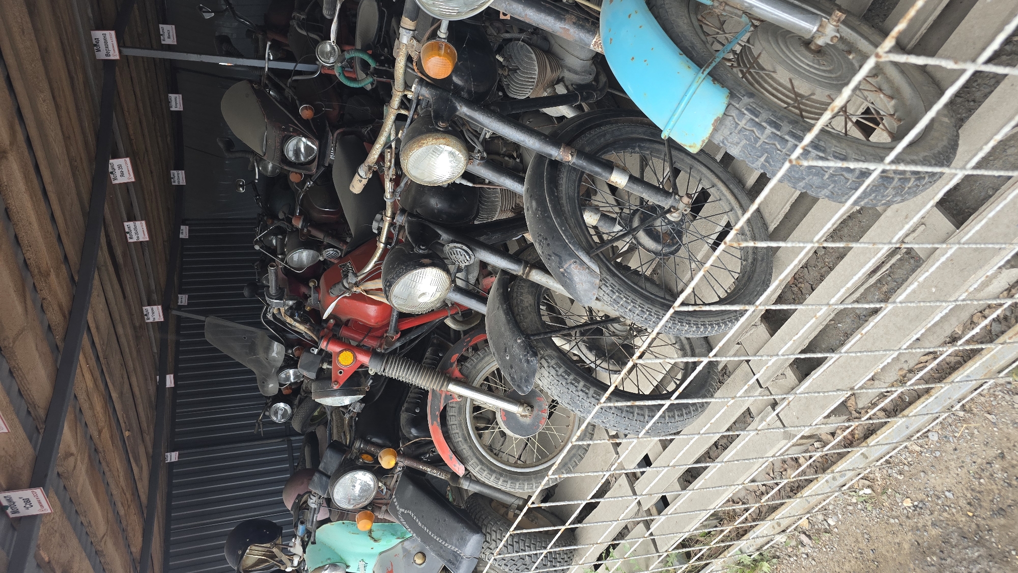 The image depicts a storage area in Bol'shoi Mashlyak, Russia, filled with vintage motorcycles. The motorcycles are lined up in rows on a wooden platform, with a wire fence in the foreground. Several of the motorcycles are red, while others are black, green, and blue. Some of the motorcycles have visible signs with Russian text, indicating their names or origins. The environment appears to be outdoors or in an open-sided structure, with a corrugated metal wall visible in the background. The lighting suggests it is daytime. There are no people visible in the image.