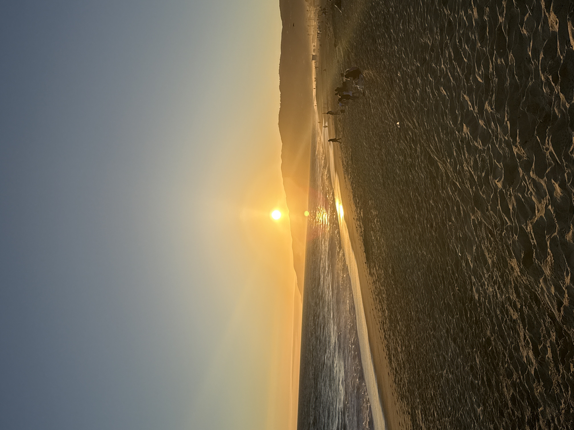 This vertical, eye-level, full shot photograph captures a sunset scene at a beach in Malibu, United States of America. The sun, a bright orb, is setting over the ocean, casting a golden glow across the sky and reflecting on the water. Gentle waves are rolling onto a sandy beach, leaving behind faint footprints. In the distance, rolling hills form a silhouetted backdrop. Several people are scattered across the beach, some walking, others sitting or standing, enjoying the view. The overall atmosphere is serene and picturesque.