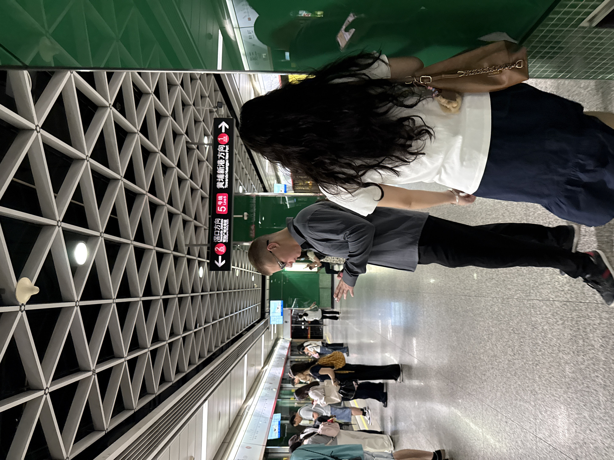 This image captures a scene on a subway platform in Tianhe District, China. In the foreground, a young man wearing a grey shirt and black pants, with glasses on, is looking down at his hands, seemingly engrossed in his phone. To his right, a young woman with long, dark, curly hair, wearing a white t-shirt and a dark blue skirt, walks away from the camera, carrying a brown leather purse with a chain strap. Behind them, a diverse group of people are standing on the platform, some looking towards the tracks, others engaged in conversation. The subway station features a modern ceiling with a geometric triangular pattern and fluorescent lighting. Signs indicating directions for subway lines are visible, with one sign pointing left for "Hu Kou Fang Xiang" (towards Hu Kou) and "Line 5", and another sign pointing right for "Huangpu Xin Gang Tou" (towards Huangpu New Port) and "Line 1". The platform floor is a light-colored speckled material, and a green wall with a textured surface is visible on the right side of the frame. The overall impression is a busy urban transit environment.