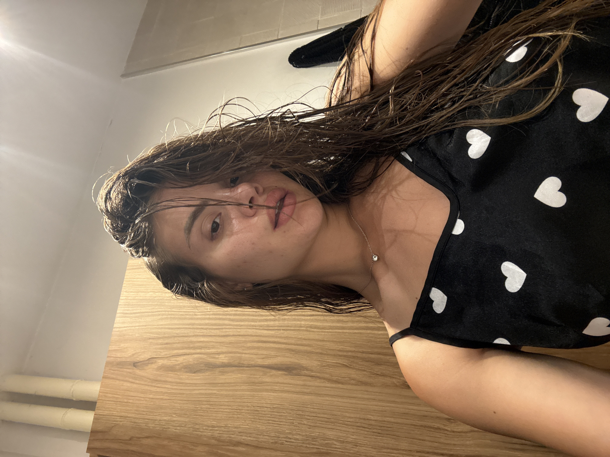The image is a rotated close-up portrait of a young woman with wet, dark brown hair. Her hair is disheveled, falling across her face and shoulders. She is wearing a black top or camisole adorned with a pattern of small white hearts. A delicate silver necklace with a small, clear pendant is visible around her neck. Her face is turned towards the camera, showing a neutral expression and natural skin with some minor blemishes.
The background includes a light-colored wooden surface, possibly a table or counter, occupying the lower portion of the rotated image. Above this, a white wall or ceiling is visible, with faint white pipes running horizontally along the wall. A bright light source creates a glare in the upper left corner of the frame. A dark, possibly black, towel or piece of clothing is partially seen behind her head. The setting appears to be indoors, suggesting a casual, personal moment, possibly after bathing, in a home environment like a bedroom or bathroom. No text is visible in the image.