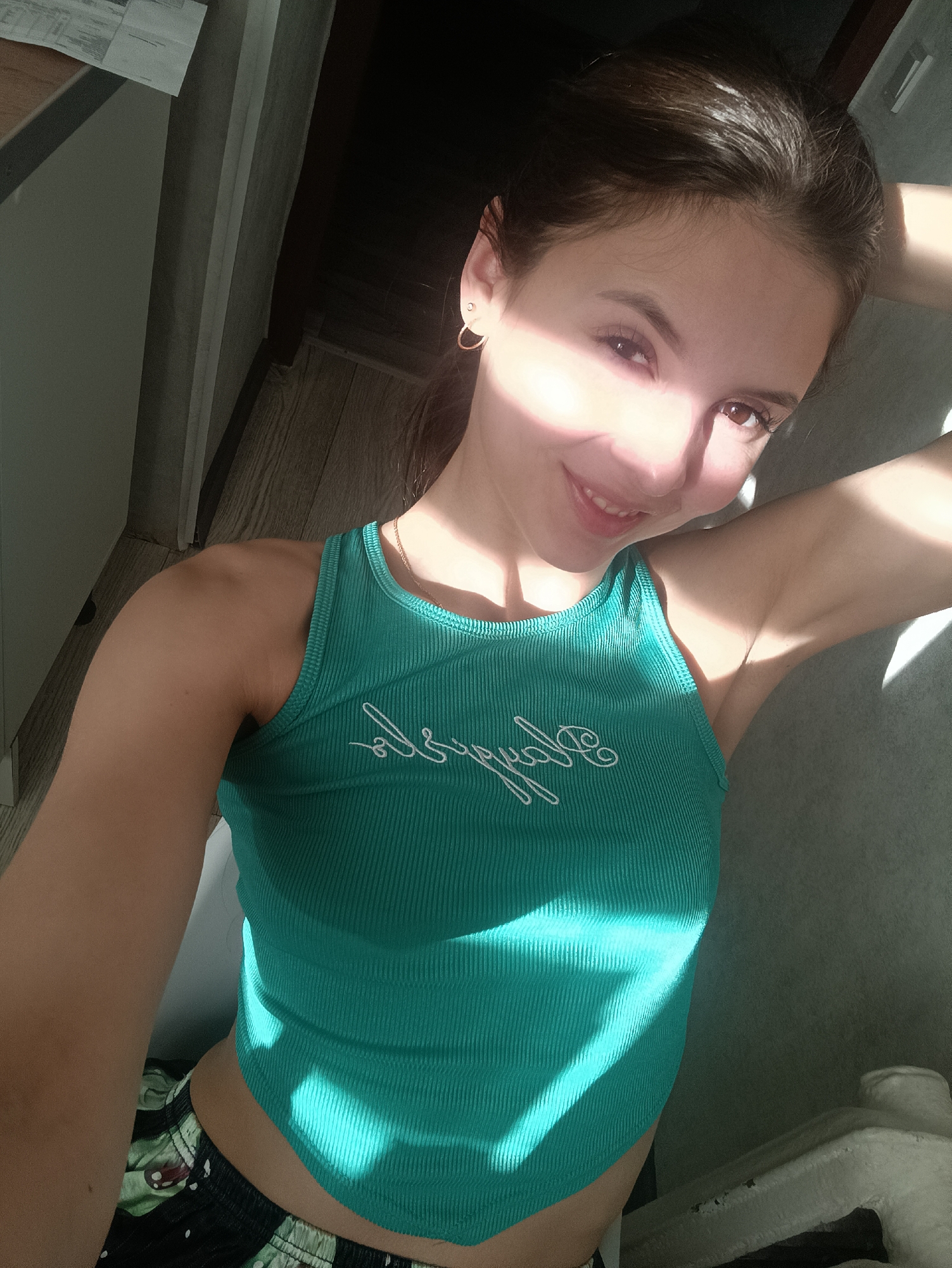 A young woman poses indoors. She has fair skin, brown hair, and is wearing a ribbed green tank top and patterned shorts. Her right arm is raised behind her head. She is smiling and looking at the camera. The background includes a wall and a door frame. Harsh sunlight is casting strong shadows on her face and body.