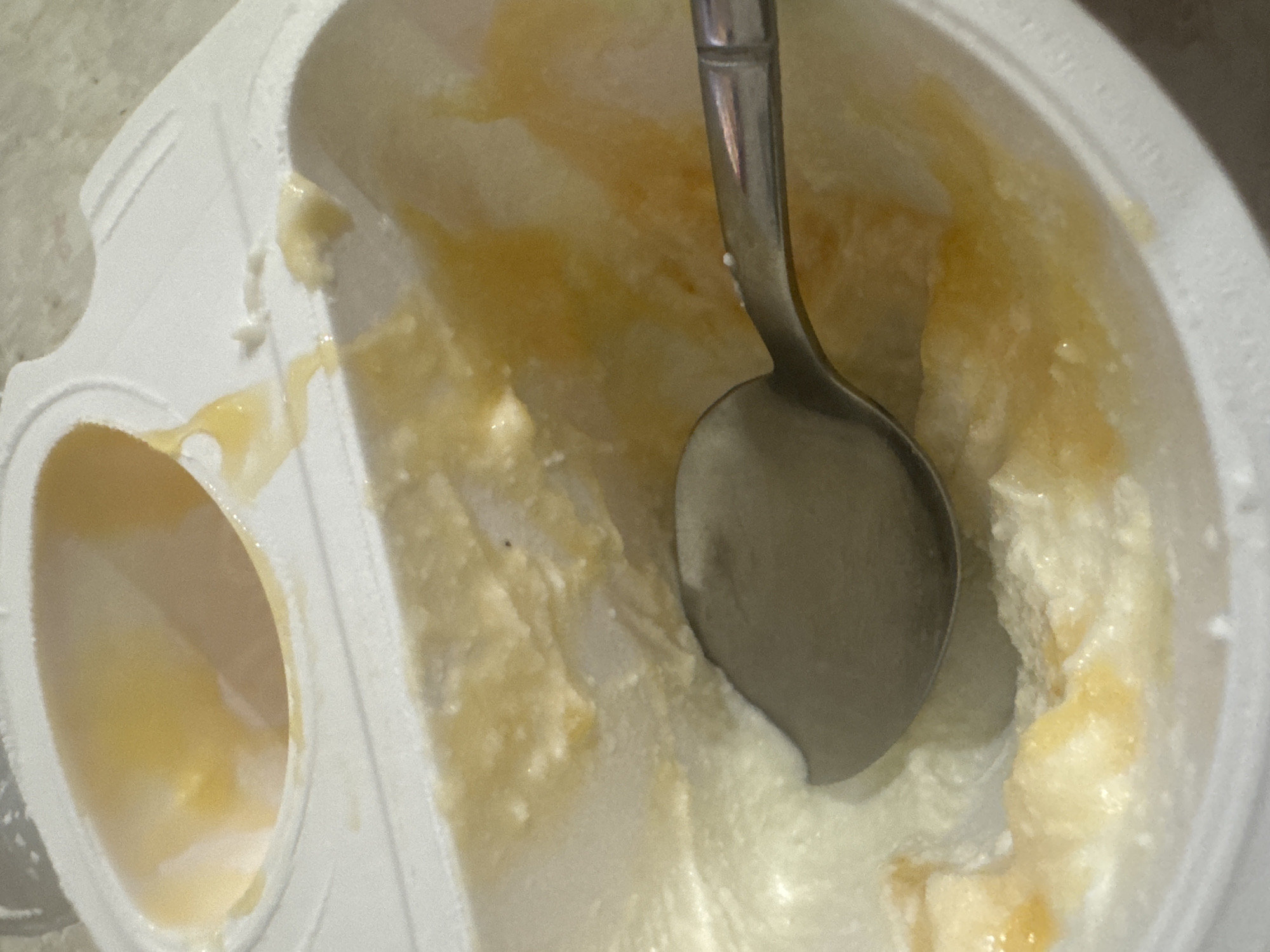 The image shows a close-up, high-angle view of a partially consumed yogurt cup. A metal spoon rests in the white yogurt, which is swirled with yellow fruit compote, likely peach or apricot. The yogurt container is white plastic and appears to be a single-serving size, with a foil lid partially removed, revealing the contents. The background is a textured, light-colored surface, possibly a countertop. The scene is indoors and there are no people visible. The image is a still life without any information that could place it in Saratov, Russia, or any specific location.