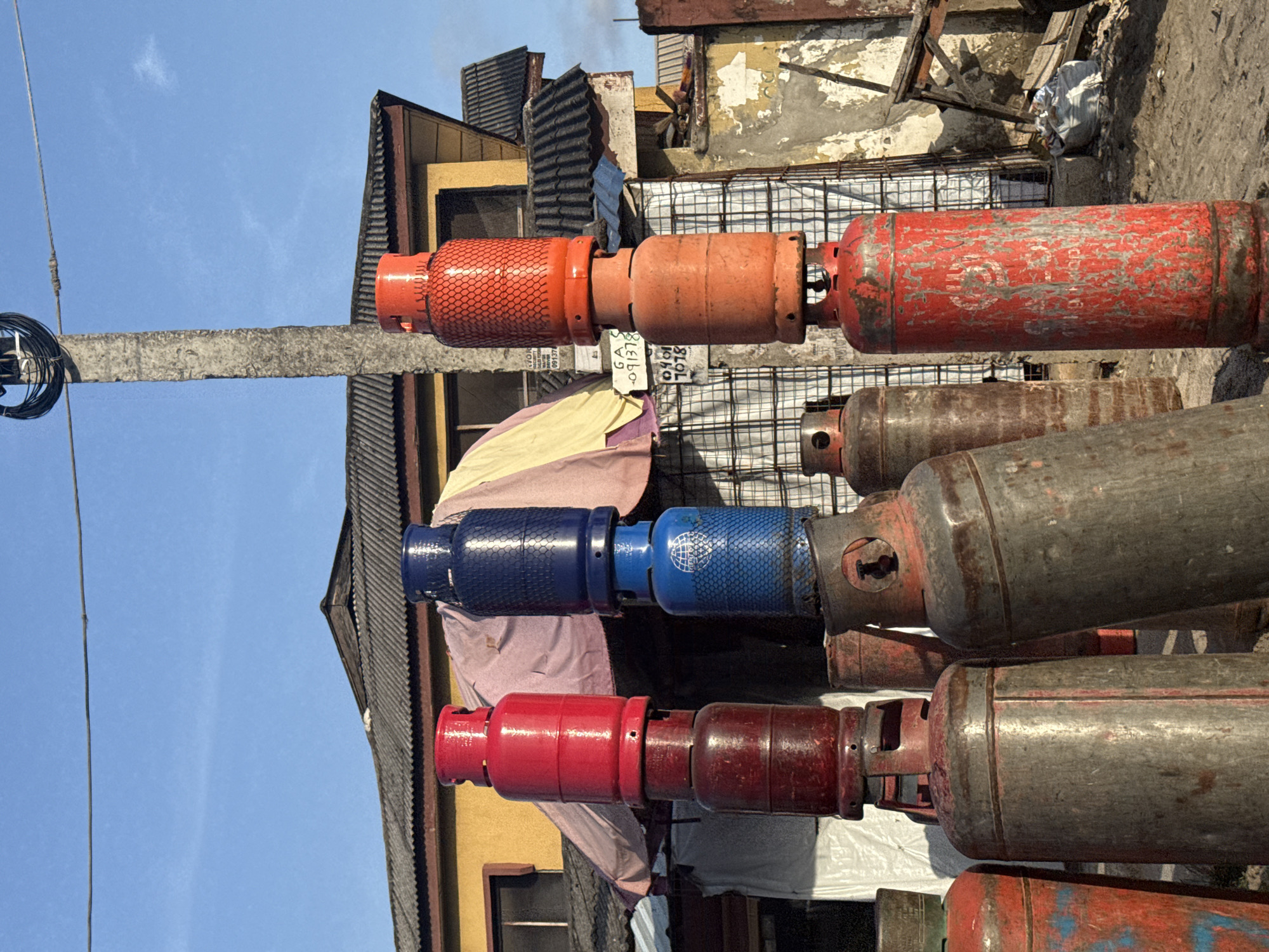 The image depicts an outdoor scene in Ado, Nigeria, characterized by a prominent display and storage of numerous liquified petroleum gas (LPG) cylinders under a clear blue sky.
In the foreground, an assortment of gas cylinders stands on sandy, uneven ground. Several are stacked vertically, with smaller cylinders balanced atop larger ones. From left to right:
* A stack features a vibrant red cylinder on top of a darker, heavily rusted red cylinder.
* Another stack displays a brightly painted blue cylinder (with a textured, netted casing) positioned above a lighter blue cylinder. The lower blue cylinder bears a visible circular logo and "GLOBAL" text.
* A third stack consists of bright orange cylinders, with the top cylinder also featuring a textured, netted casing.
* Numerous other cylinders of varying sizes, predominantly metallic grey with patches of faded red or blue paint and significant rust, are scattered across the ground. Some show extreme wear, with paint peeling extensively to reveal the underlying metal.
In the mid-ground, a tall, grey concrete utility pole, heavily textured, rises centrally. It supports a cluster of tightly wound black cables near its top and a horizontal wire stretching across the upper frame. Behind the cylinders, a yellow building with a dark corrugated metal roof is visible. An awning, striped in yellow and purple/pink hues, extends from this building. Further right, parts of another building with a similar corrugated roof and heavily distressed, peeling walls are present. A white sign with black text, including "GA -091378" and "0901 7078," is attached to a metal grid structure in the background. Debris, including a white sack and wooden fragments, is scattered on the ground near the distressed wall.
No individuals are visible within the frame. The overall impression is that of a commercial gas refilling or distribution point.